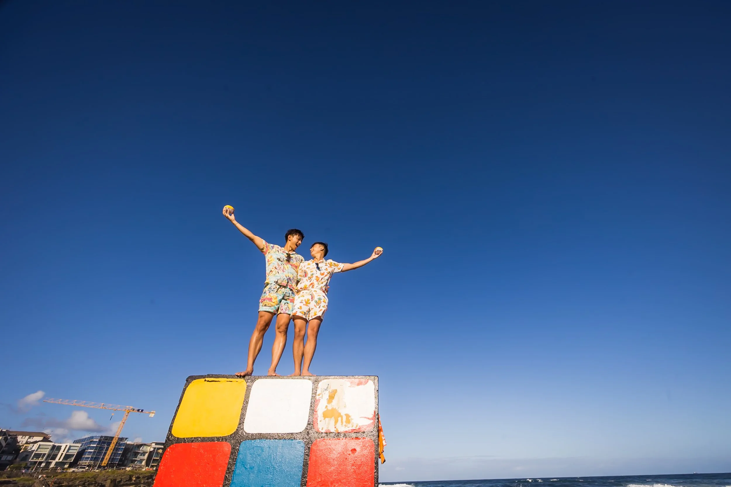 Two people standing on top of a colorful Rubik's cube sculpture at the beach, holding small balls and celebrating under a clear blue sky. Maroubra Beach  Sydney - Katsu Nojiri Sydney Wedding Photographer