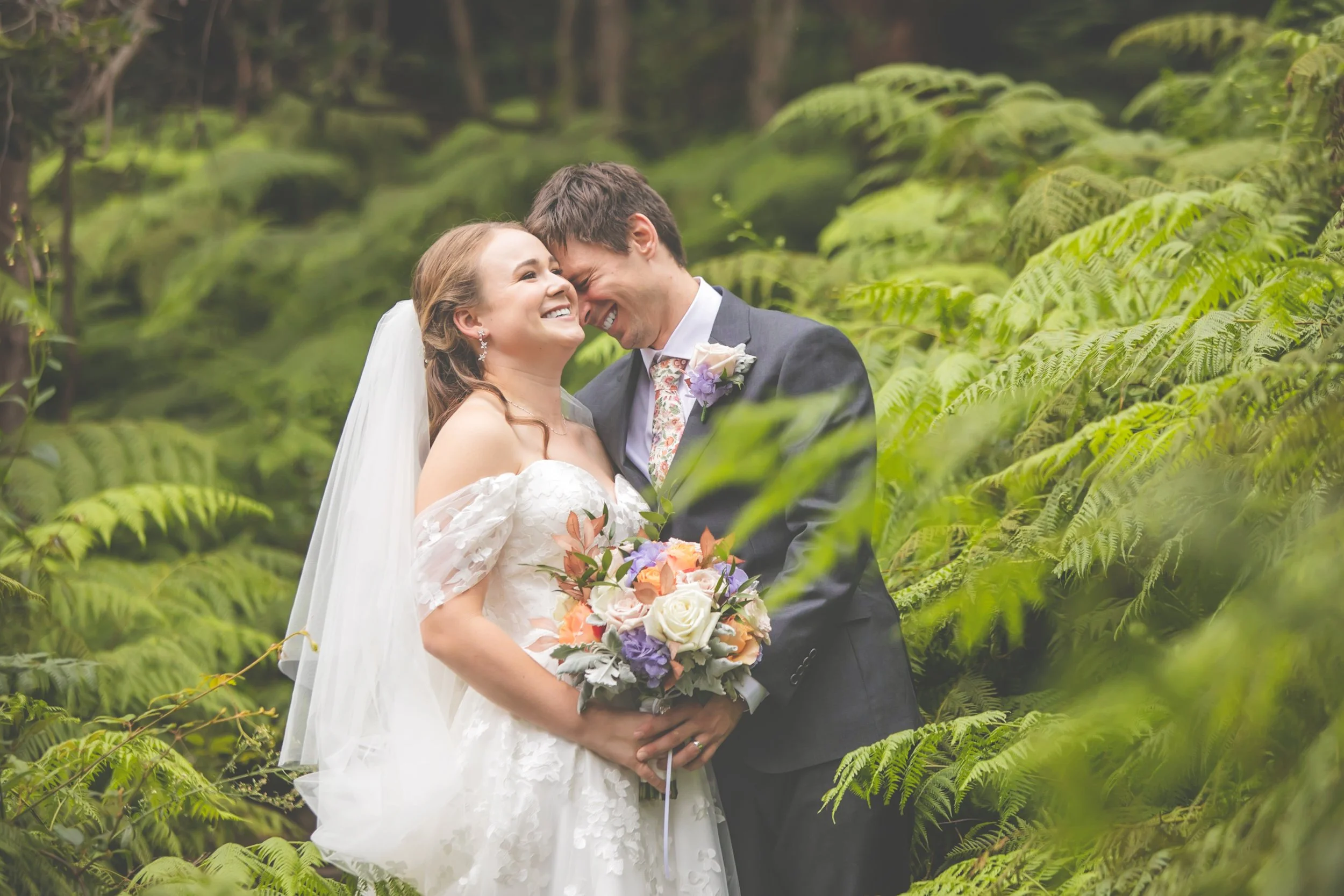 A bride and groom on their wedding day, embracing in a lush green outdoor setting, smiling and holding a bouquet of flowers at Sergeants Mess