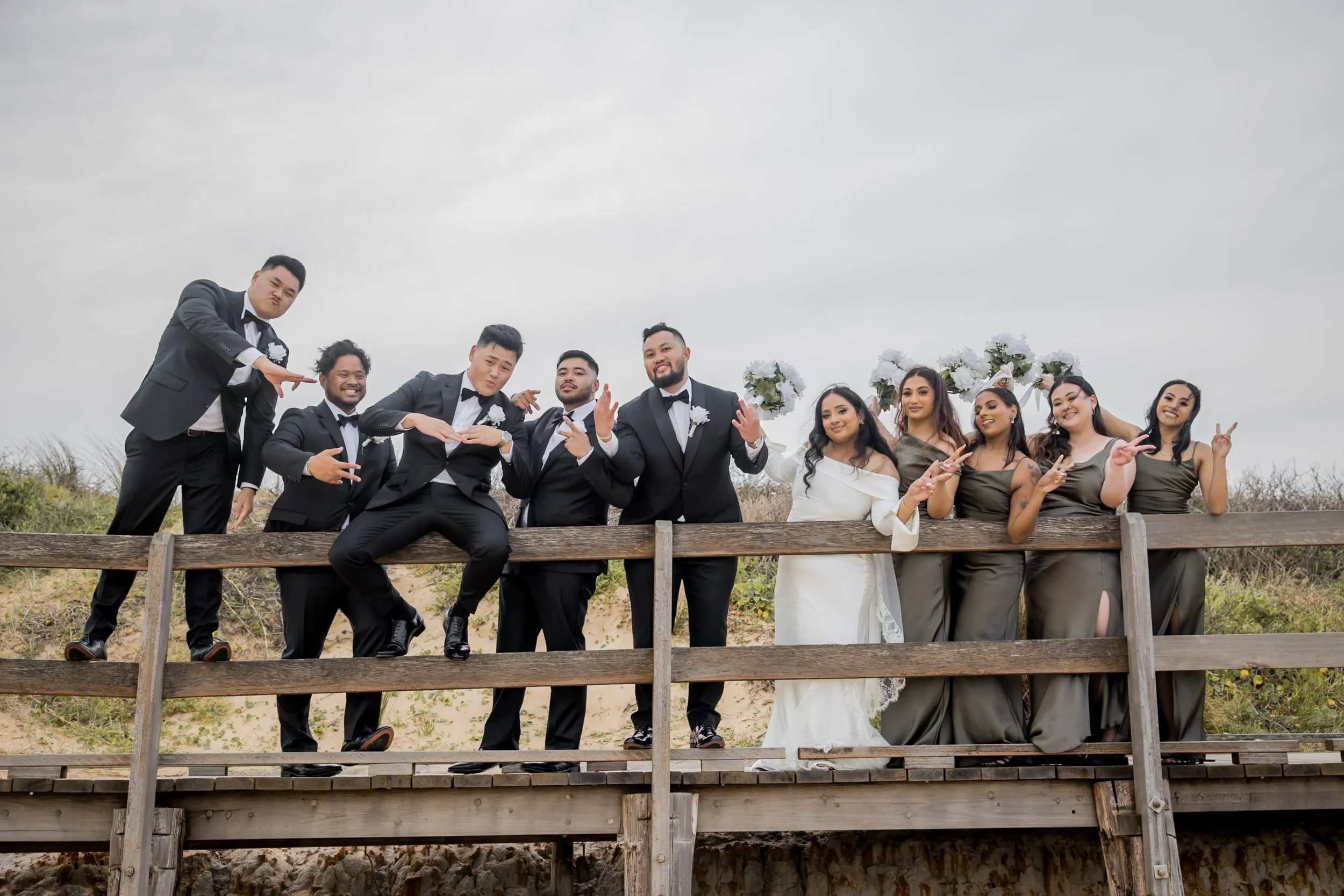 A wedding party of ten people, including the bride and groom, posing on a wooden bridge outdoors. The group includes five men in tuxedos and five women in matching dresses, . - Long Reef Golf Course  - Katsu Nojiri Sydney Wedding