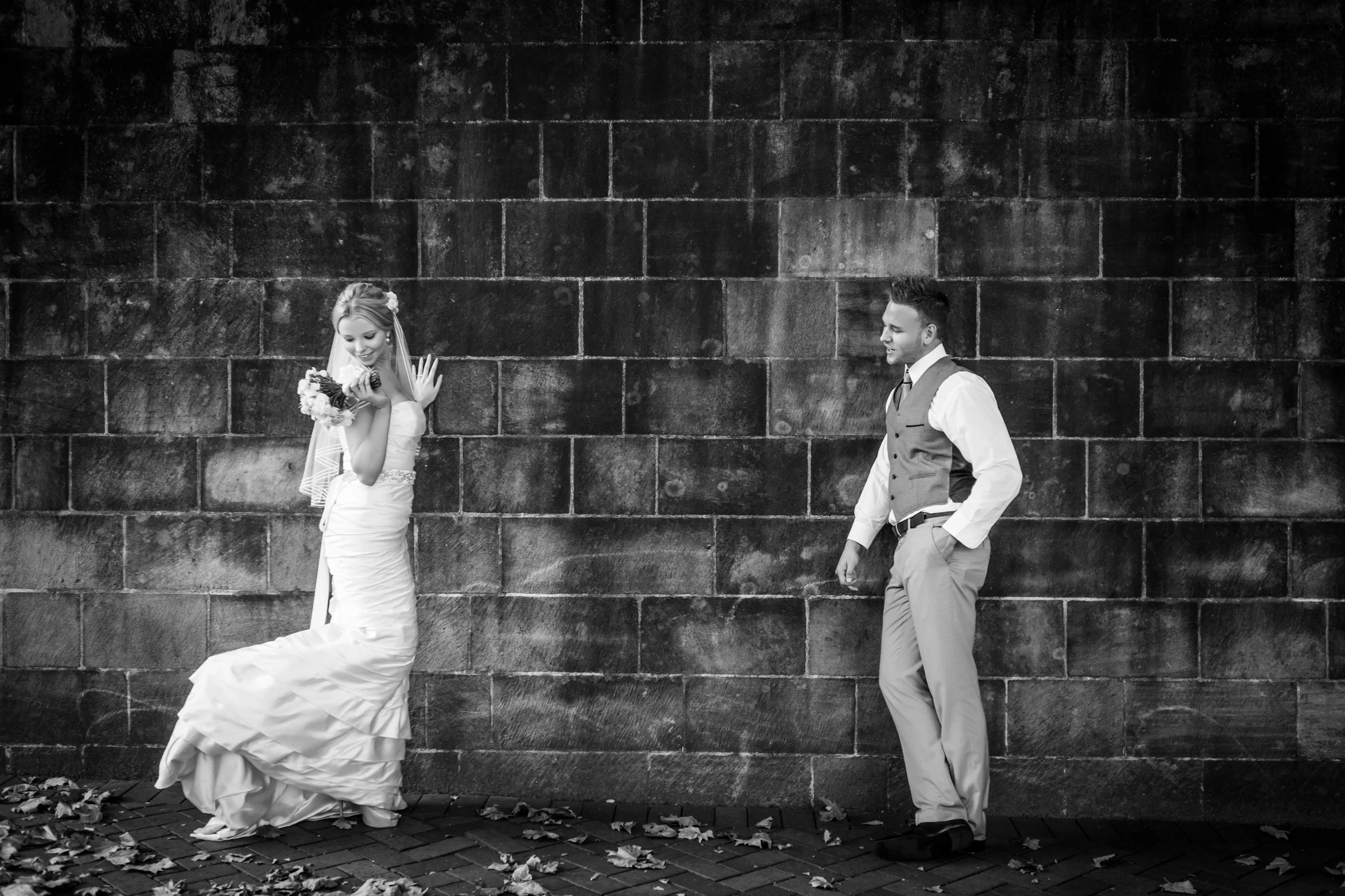 Black and white photo of a bride in a wedding dress holding a bouquet, smiling and looking down, while a groom in a suit with a vest watches her, standing against a brick wall.