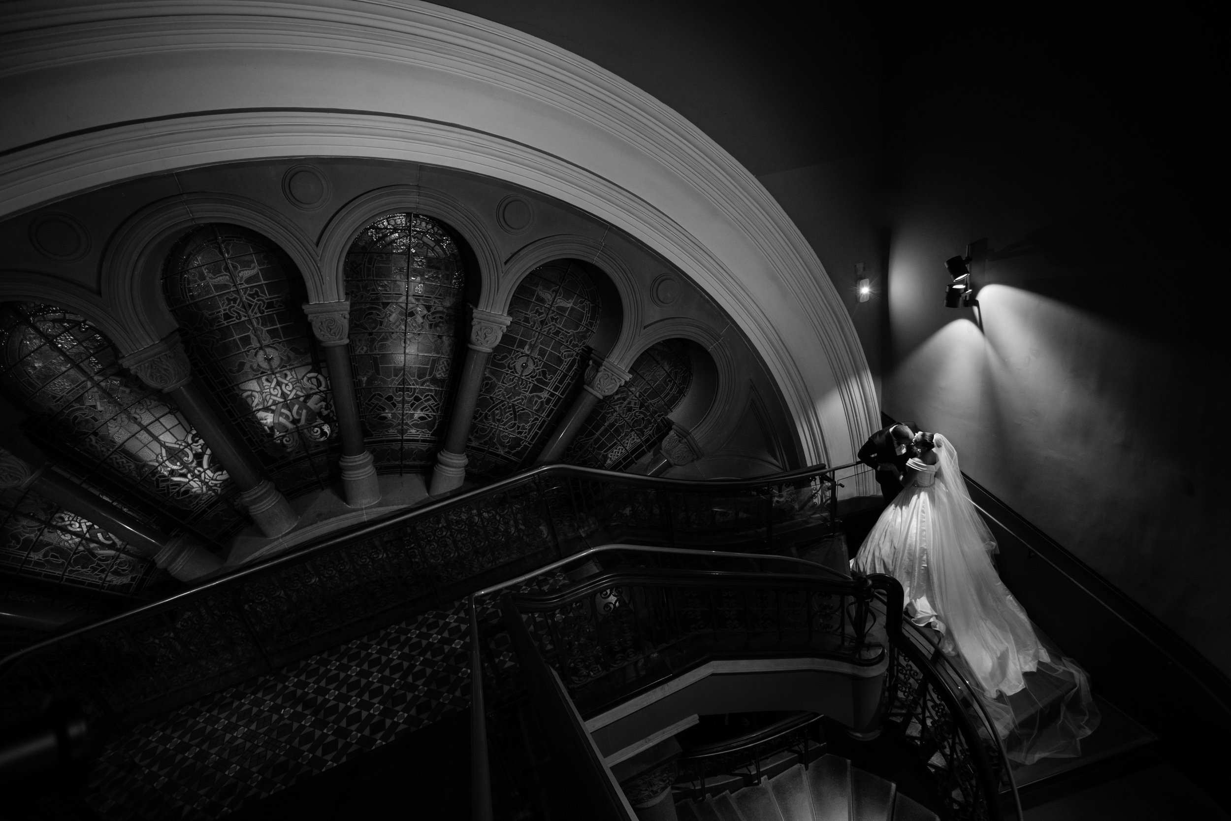 Black and white photograph of a bride and groom sharing a kiss on a grand staircase with ornate ironwork, stained glass windows, and dramatic lighting.
