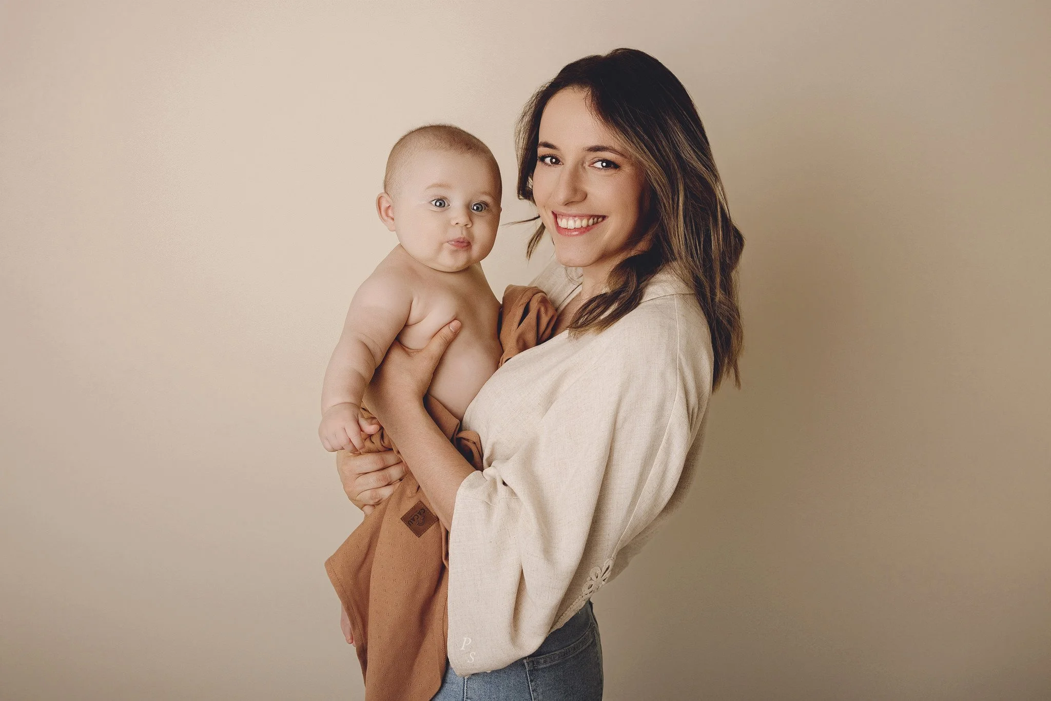 Mãe sorridente segurando um bebê de colo, com fundo bege  sessão fotográfica em estúdio no Porto