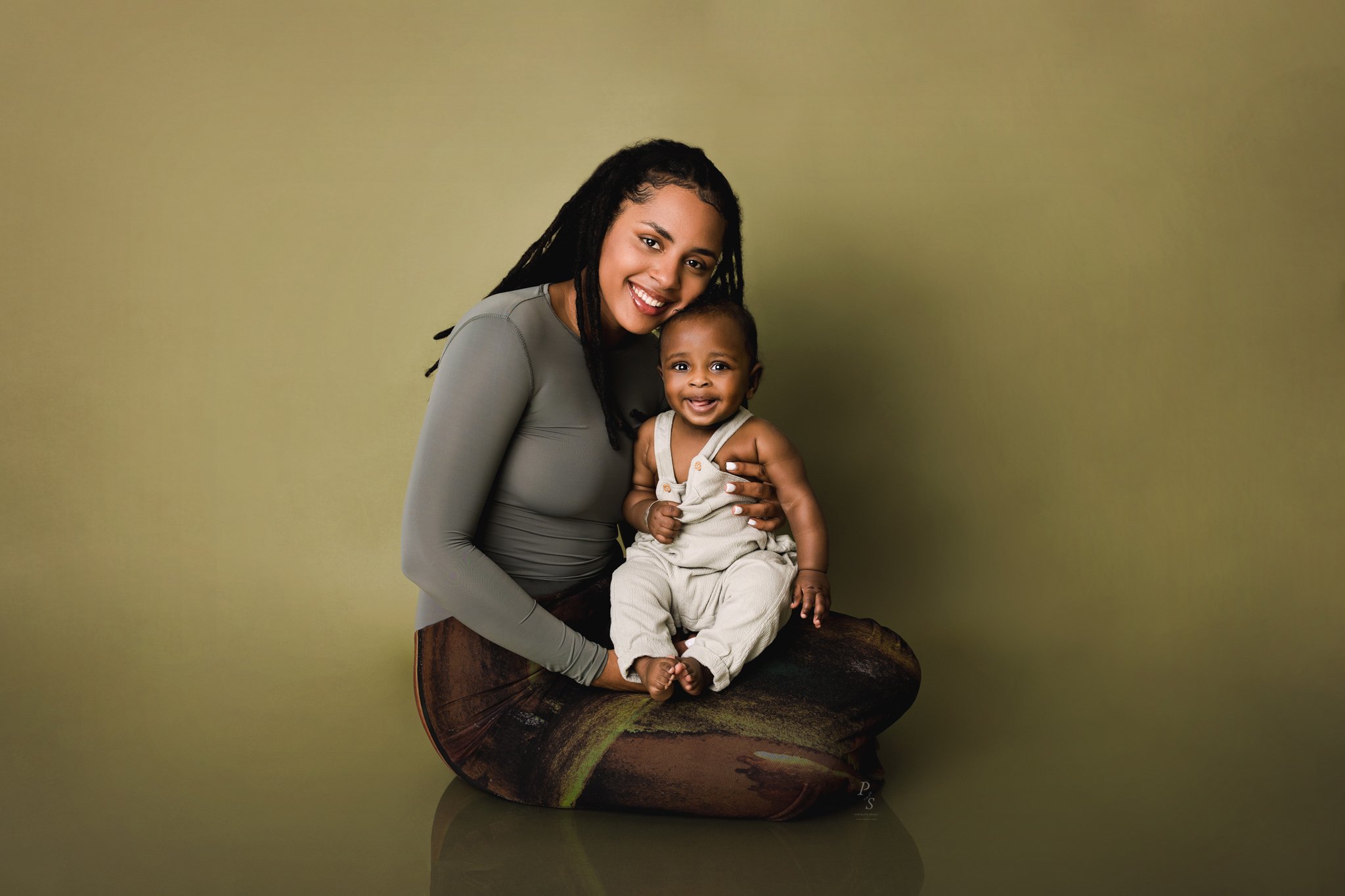 Mulher sorridente a segurar um bebé num fundo verde oliva, ambos a olhar para a câmara. sessão de família em estúdio fotográfico no Porto.