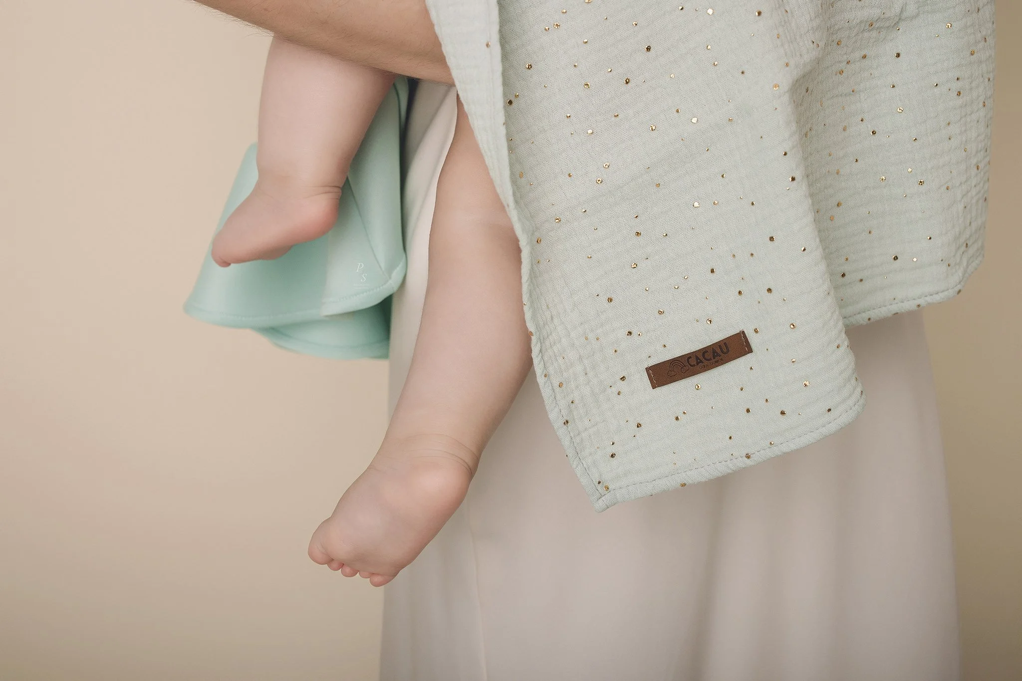 Detalhe de uma criança sendo carregada por uma pessoa, mostrando a mãozinha do bebê segurando a roupa da pessoa que o segura. Roupas de cores claras, uma com detalhes de pequenas lantejoulas douradas e uma etiqueta de couro com a marca 'Cacau'.  