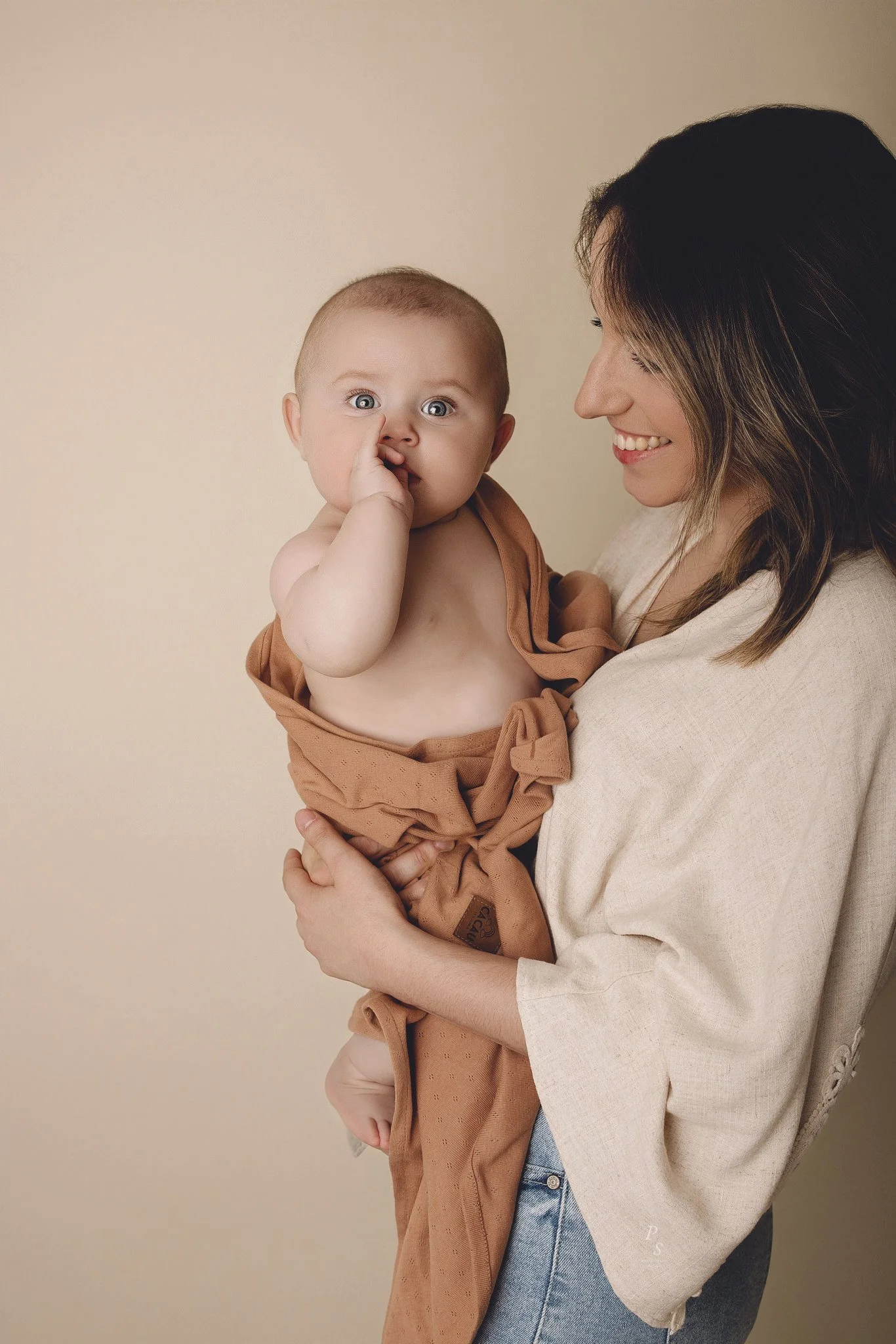 Mãe sorridente segurando um bebê com pele clara e cabelo curto, o bebê está com o dedo na boca, contra um fundo bege.  sessão fotográfica em estúdio no Porto