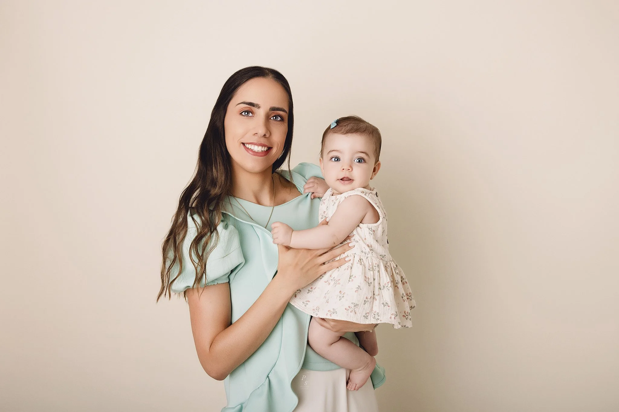 Mulher sorridente segurando uma criança pequena, ambas estão de frente para a câmara, com fundo claro e neutro.  sessão fotográfica em estúdio no Porto