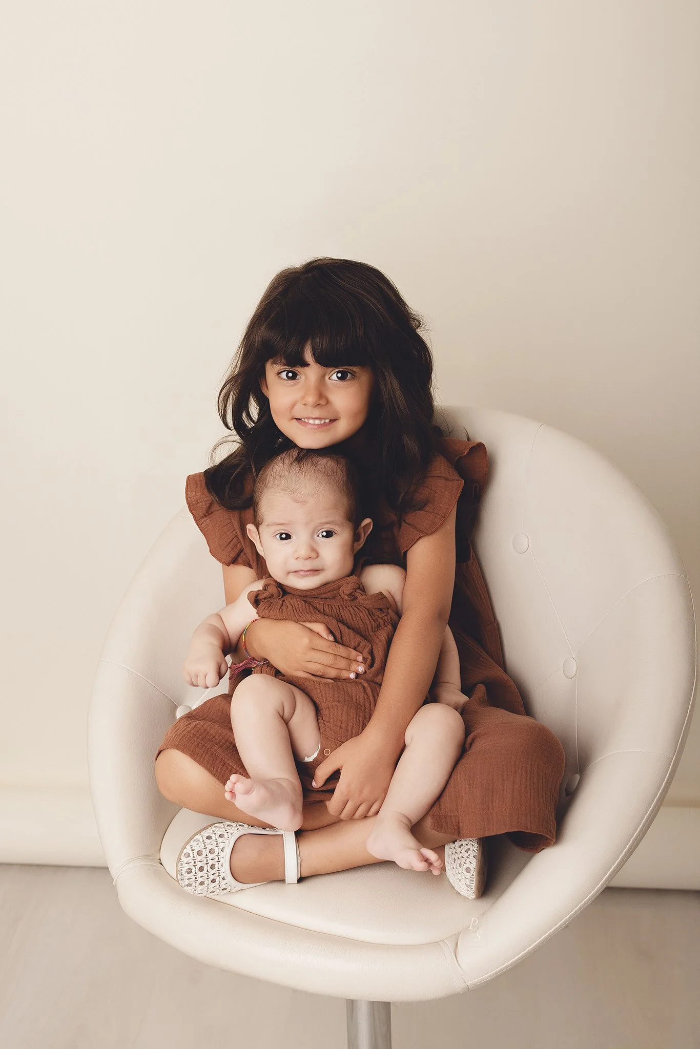 Duas crianças, uma mais velha com cabelo castanho escuro e cacheado, sorrindo, segurando uma mais nova com cabelo liso e mais clara, ambas vestindo roupas de tom castanho, sentadas em uma cadeira branca.  sessão fotográfica em estúdio no Porto
