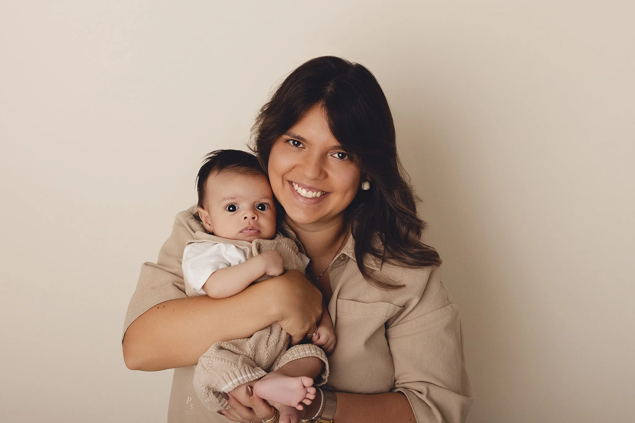 Mulher sorridente segurando um bebê, ambos posando contra fundo bege.  sessão fotográfica em estúdio no Porto