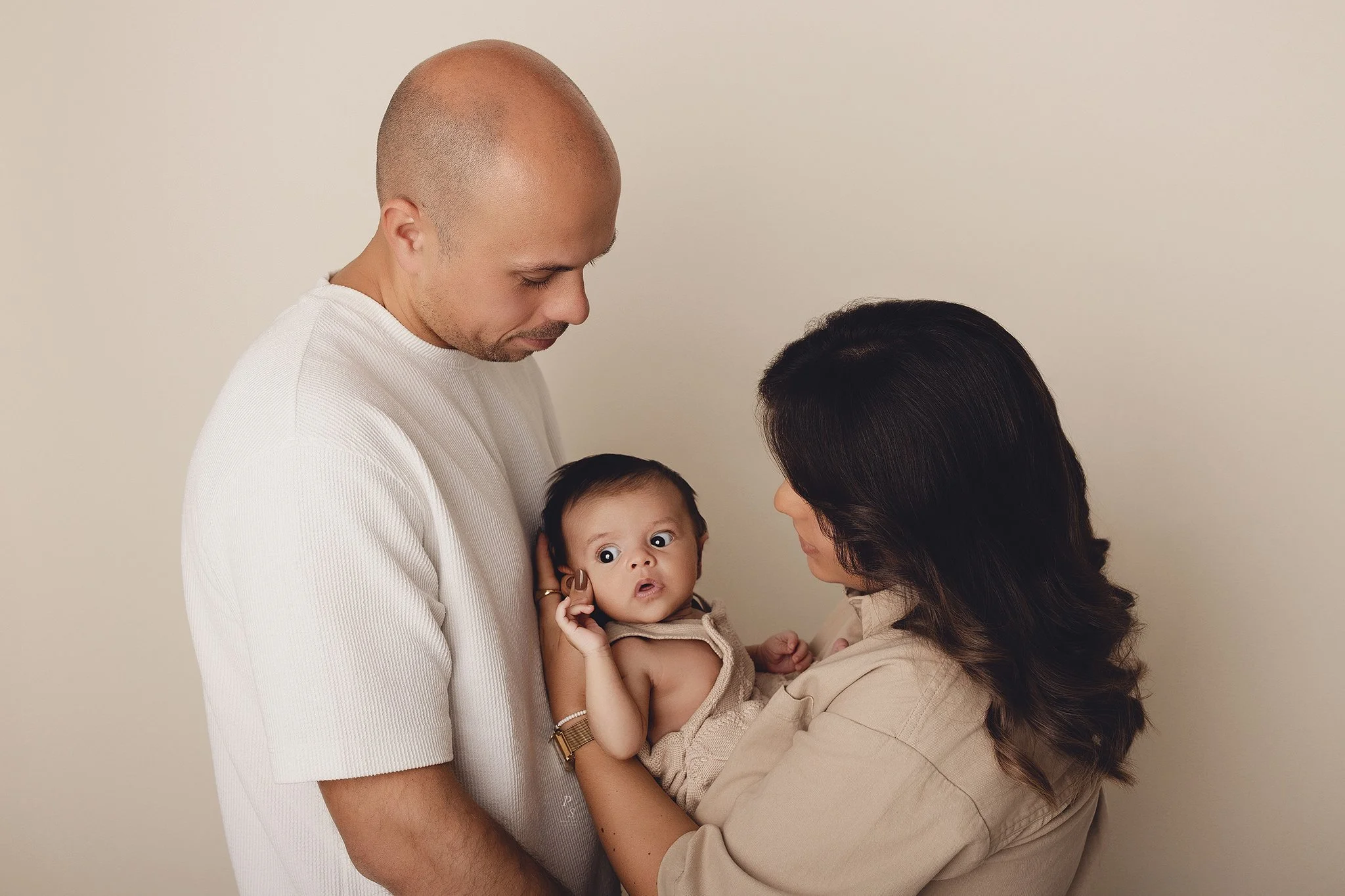 Uma família de três pessoas, mãe, pai e bebé, juntos em um ambiente interno com fundo bege. A mulher segura o bebê, que olha para a câmera com expressão surpresa, enquanto o homem observa afavelmente.  sessão fotográfica no porto