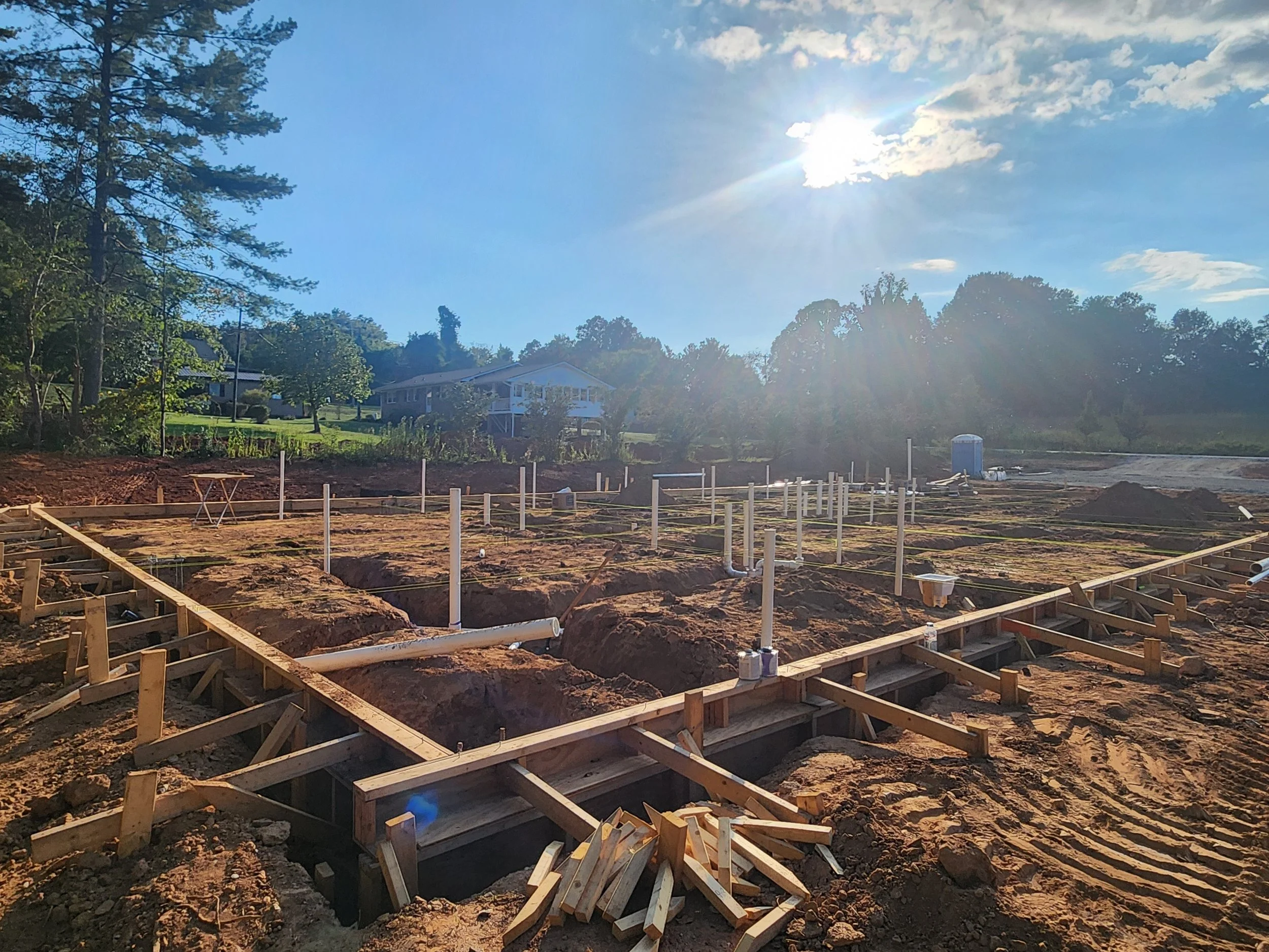 Construction site with wooden framework and pipes for a building foundation, under a bright sky with the sun shining.