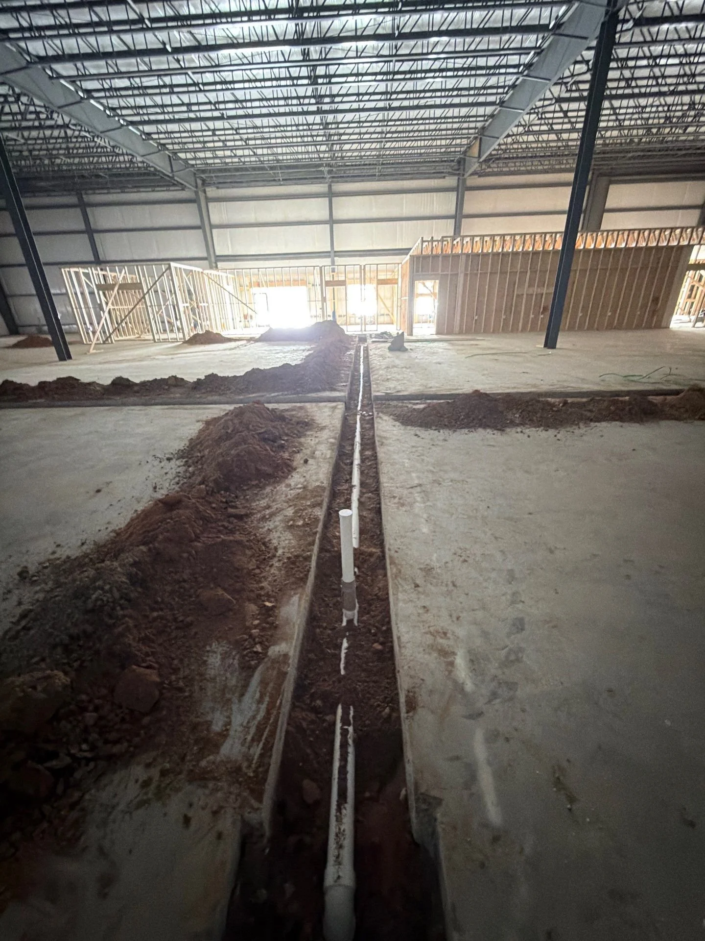 Interior of a building under construction with exposed steel framework, partially built wooden structures, and an open trench with PVC pipes in the center, sunlight coming through the openings.