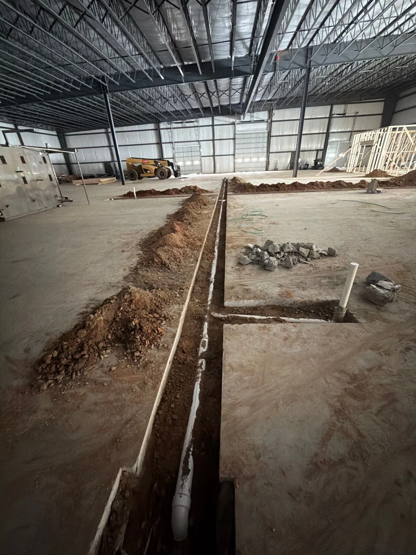 Construction site inside a large building with exposed steel framing, ongoing electrical or plumbing work with a long trench marked for utility installation, construction materials, and equipment in the background.