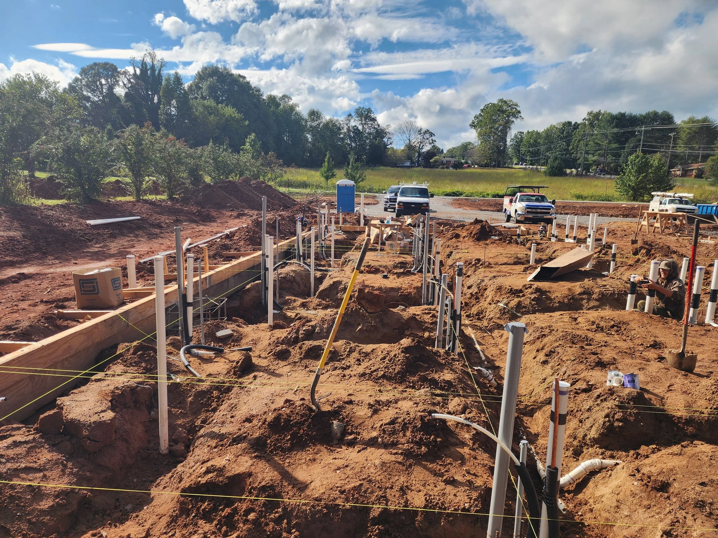 Construction site with workers installing underground pipes, multiple white pipes protruding from the ground, and vehicles parked in the background under a partly cloudy sky.