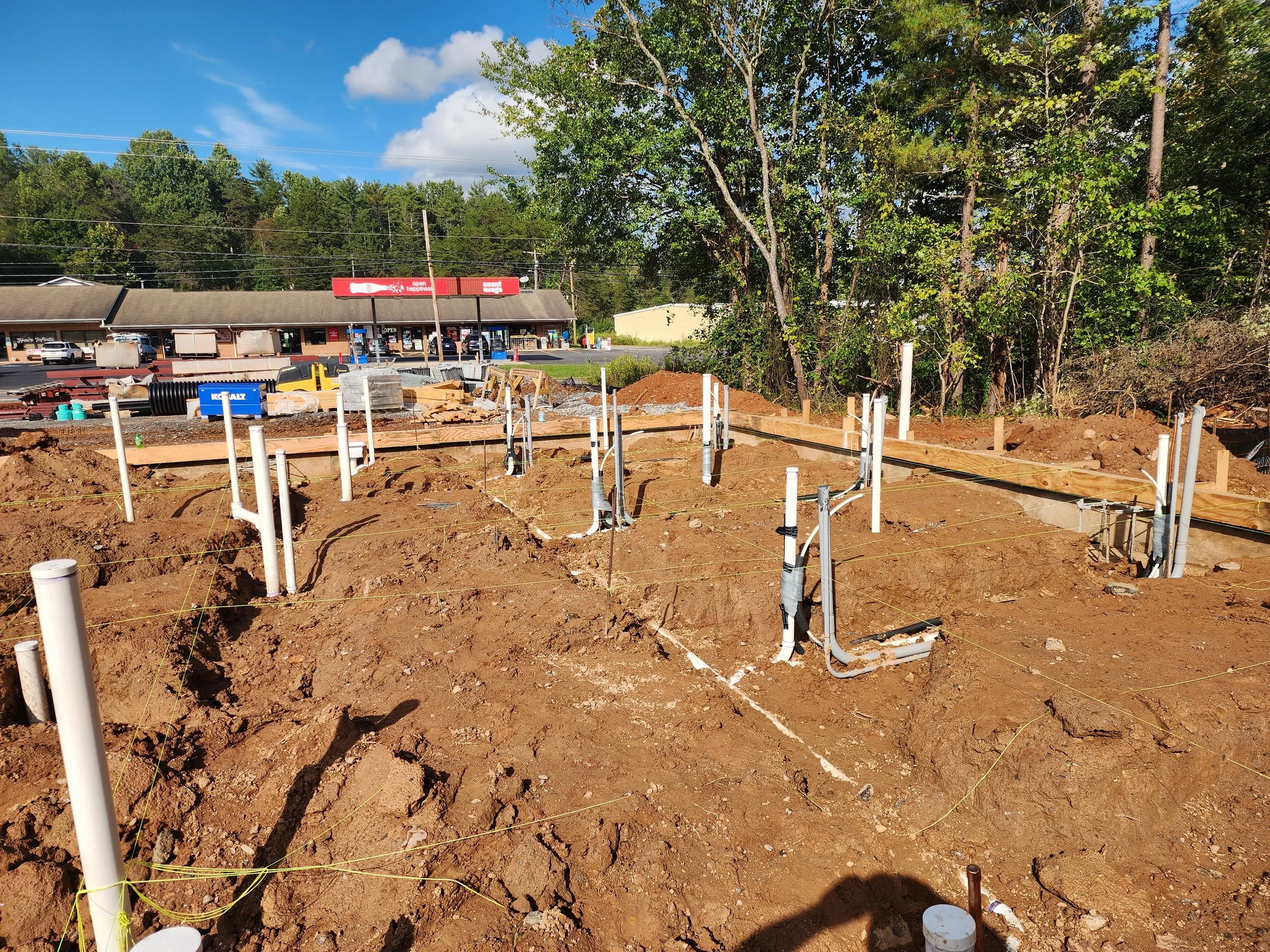 Construction site with exposed pipes and wooden framing in the foreground, a strip mall with a Redbox kiosk in the background, and trees under a partly cloudy sky.