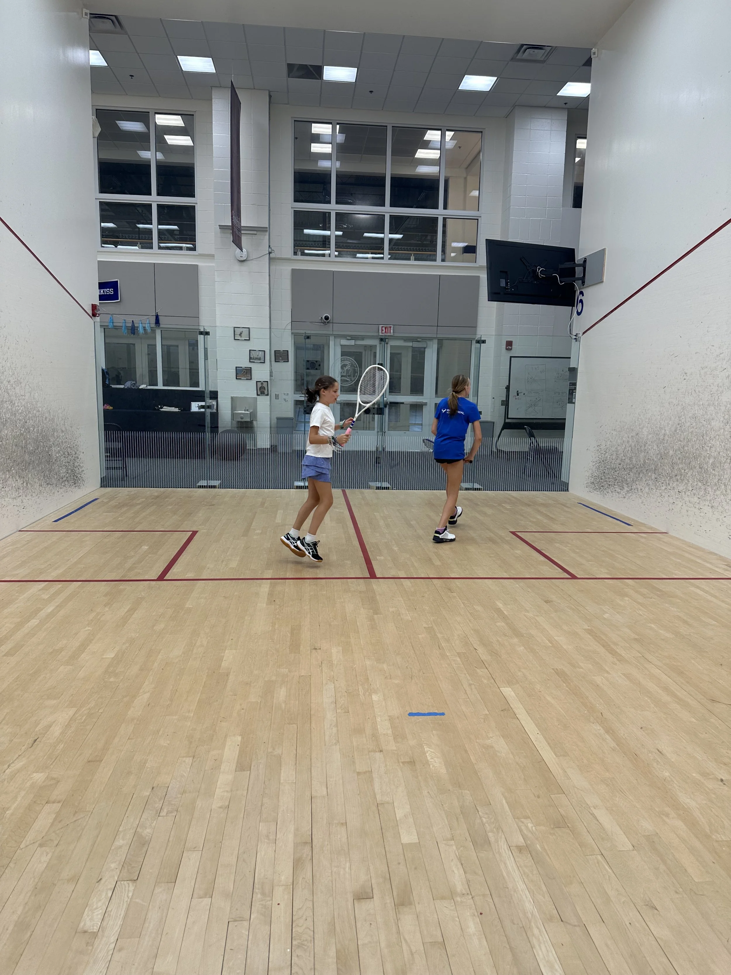 Two young girls practice on a squash court, one holding a racket and preparing for a shot while the other moves toward the back wall.