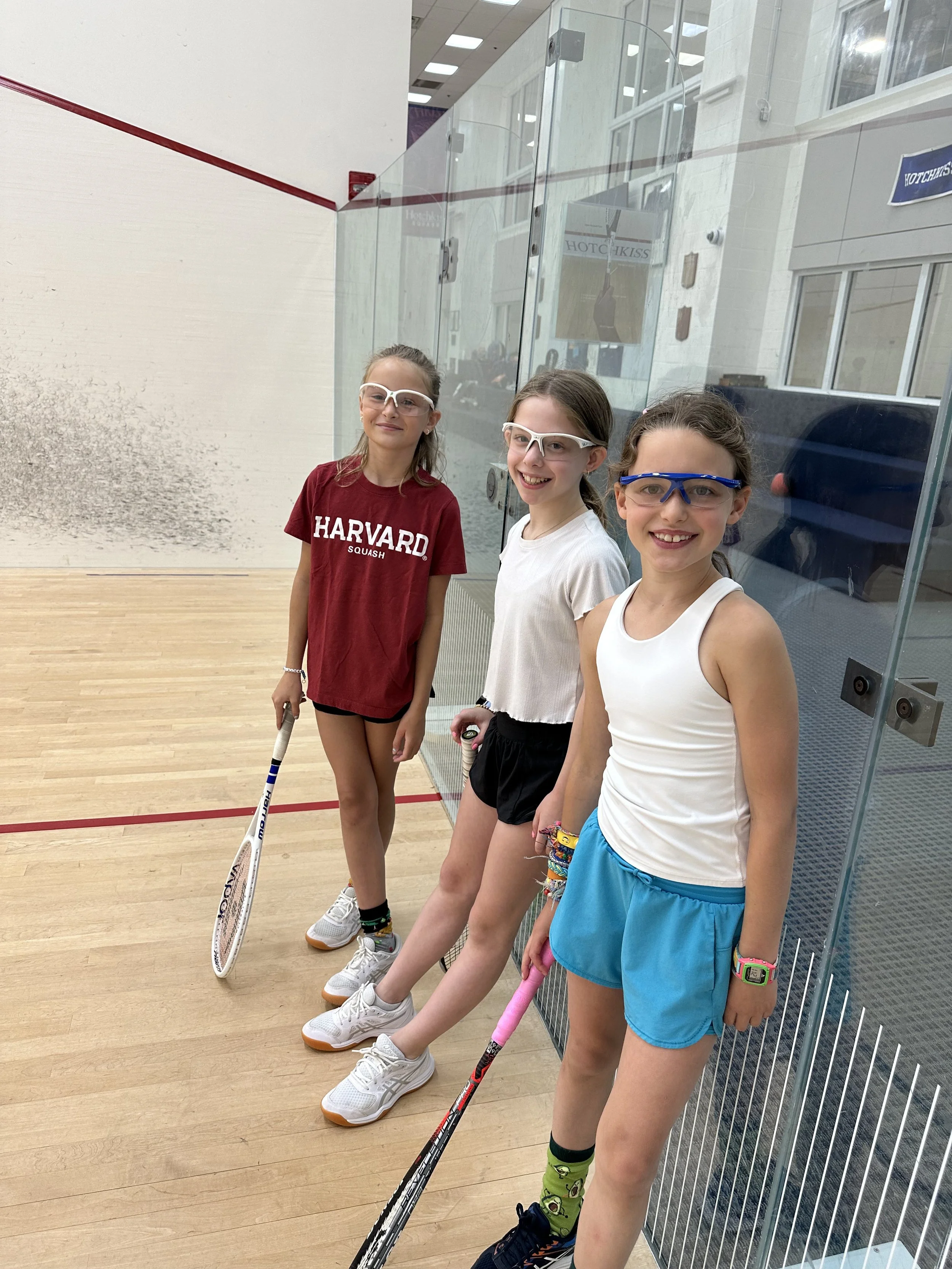 Three young girls stand on a squash court, wearing goggles and holding rackets, smiling brightly after a game or practice.