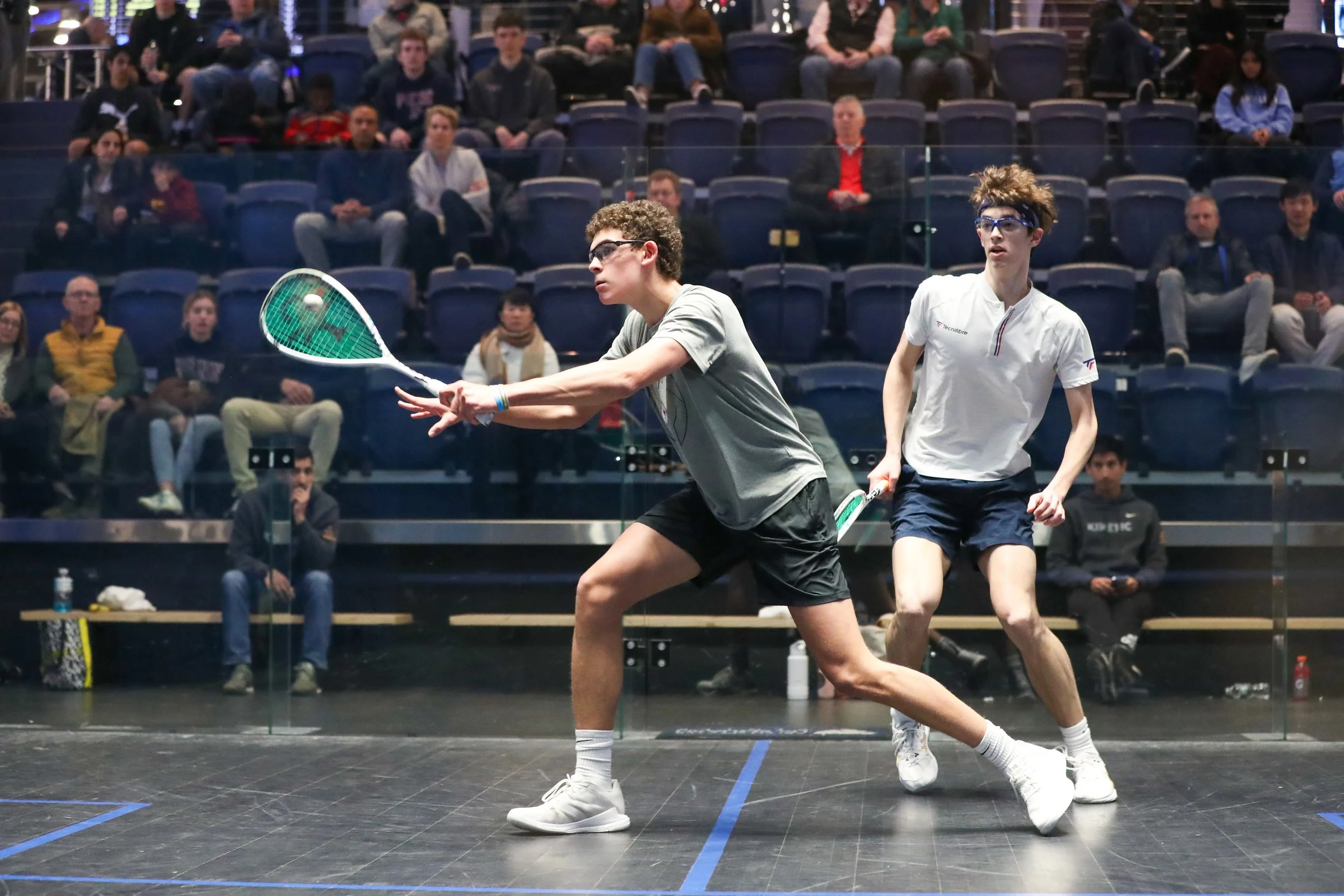 Two squash players in action during a match on a glass court, with a focused audience seated in the background.