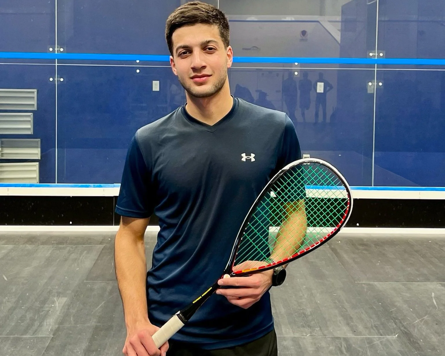A squash player holding a racket, standing confidently in front of a glass squash court.
