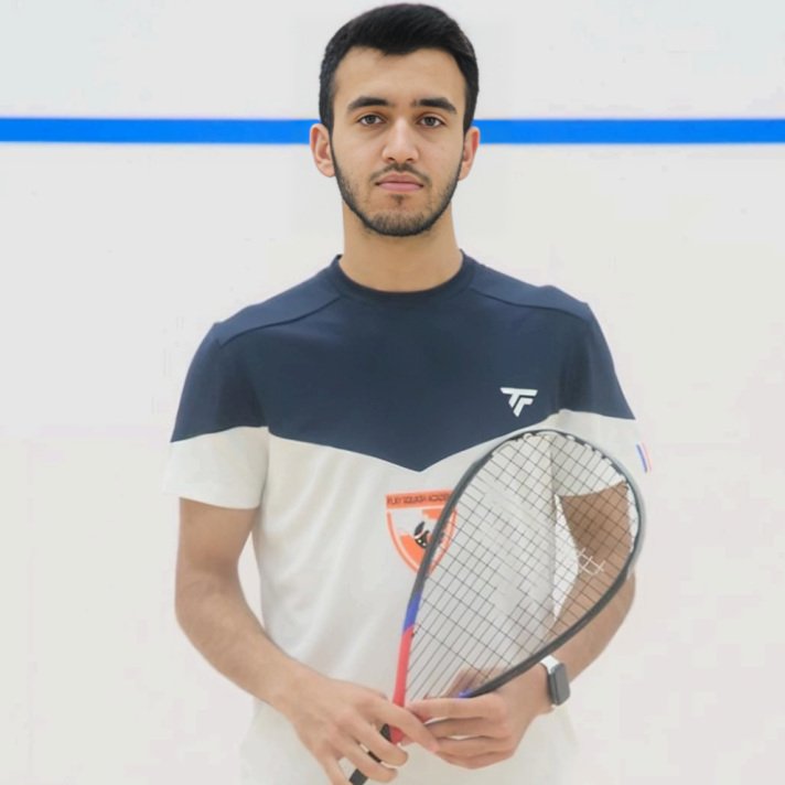 A focused individual holding a squash racket, wearing a navy and white sports shirt, posed against a clean squash court background.