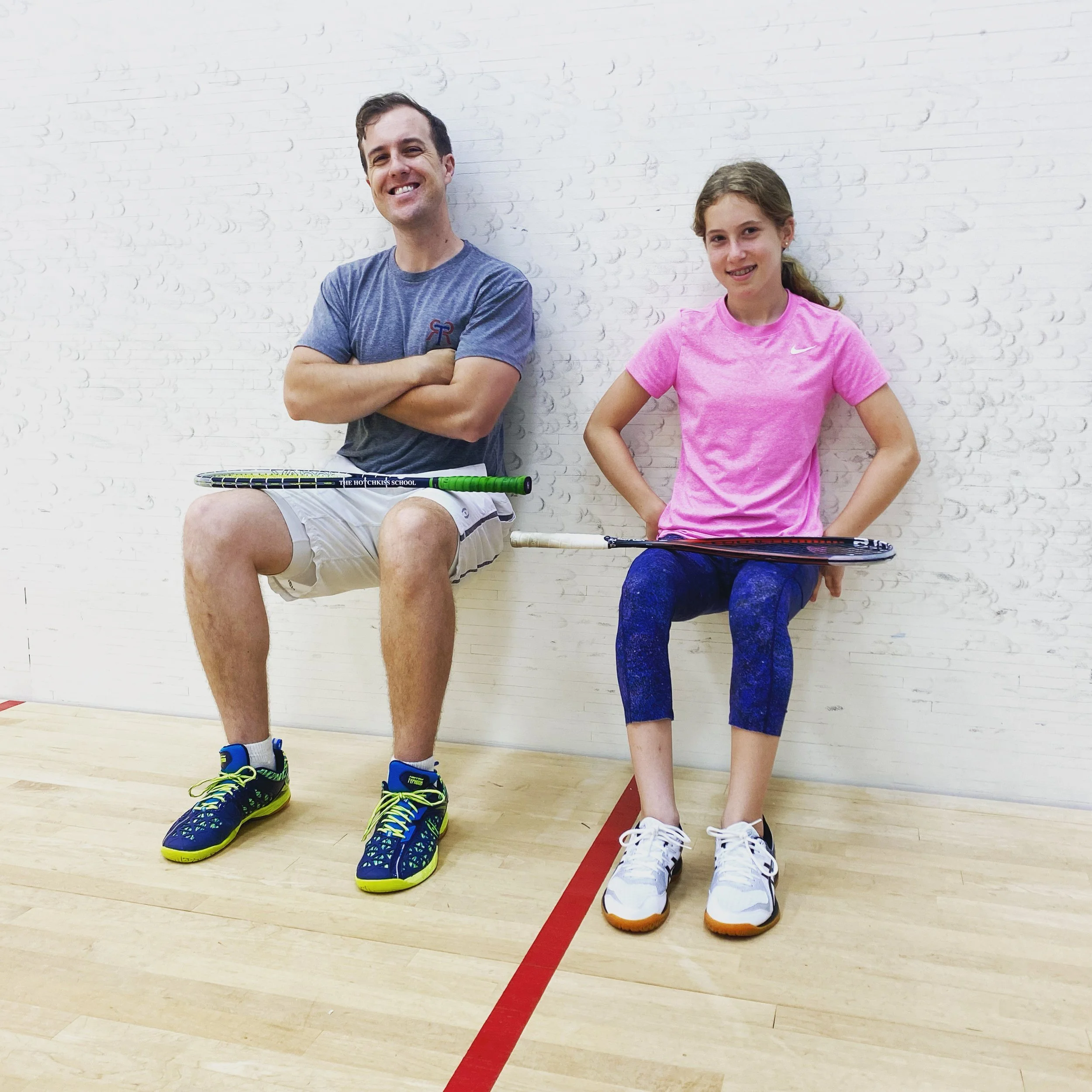 An adult and a young girl do wall sits on a squash court, smiling and balancing rackets on their laps during a fitness training session.