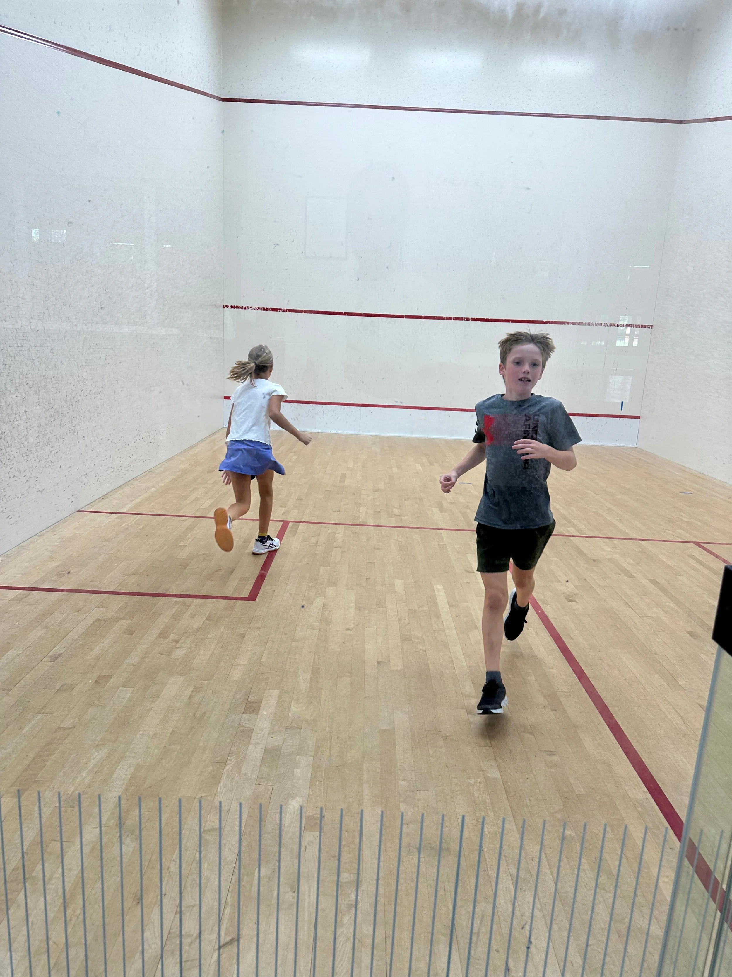 Two children run across a squash court during a fun drill or practice session, showcasing energy and enthusiasm for the sport.