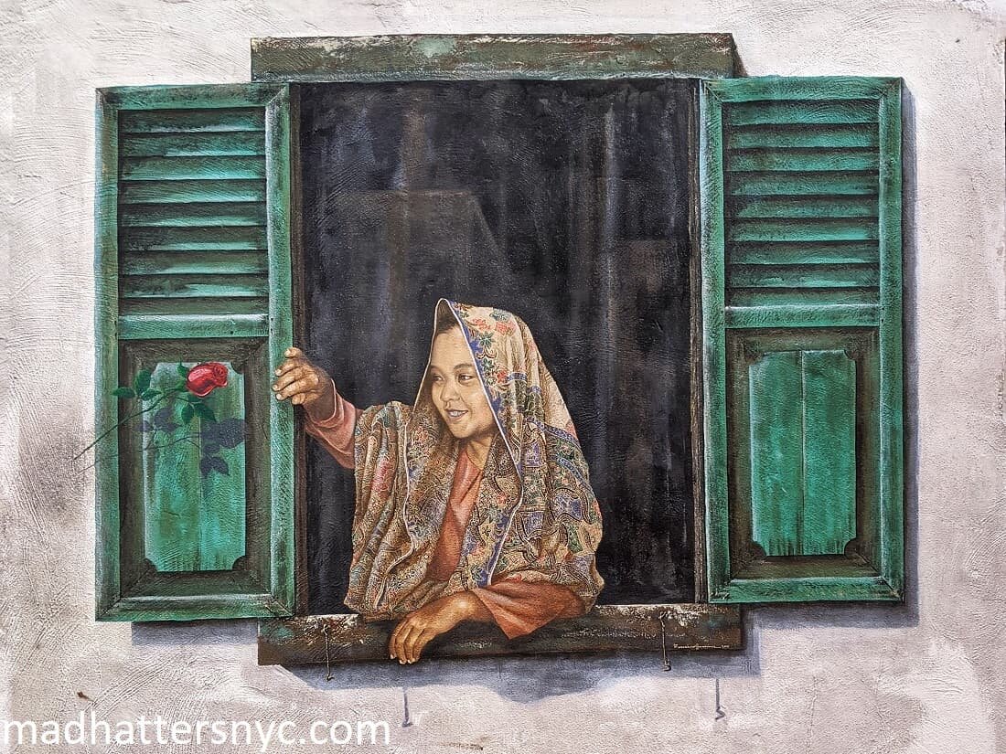 Mural featuring a girl at her louvered window wearing a batik scarf over her hair and smiling at a rose in Melaka, Malaysia