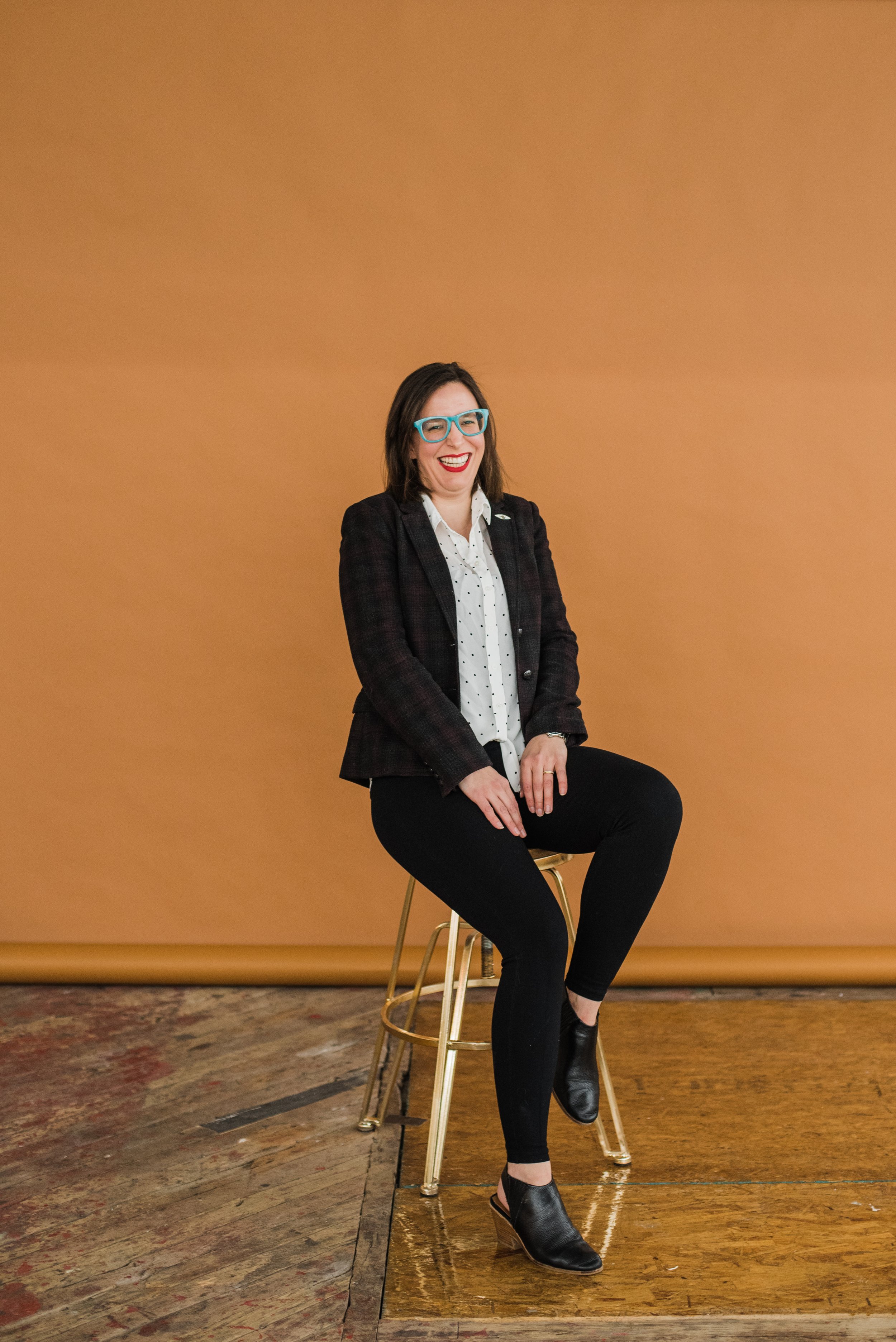Elle, sitting on a stool in front of a warm orange background. She is wearing a blazer and black pants and leaning back, laughing.