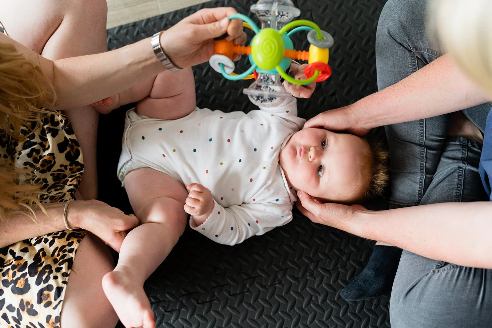 Baby lying on a black padded surface, surrounded by three adults, with one holding a colorful toy above the baby, and another gently supporting the baby's head with both hands.