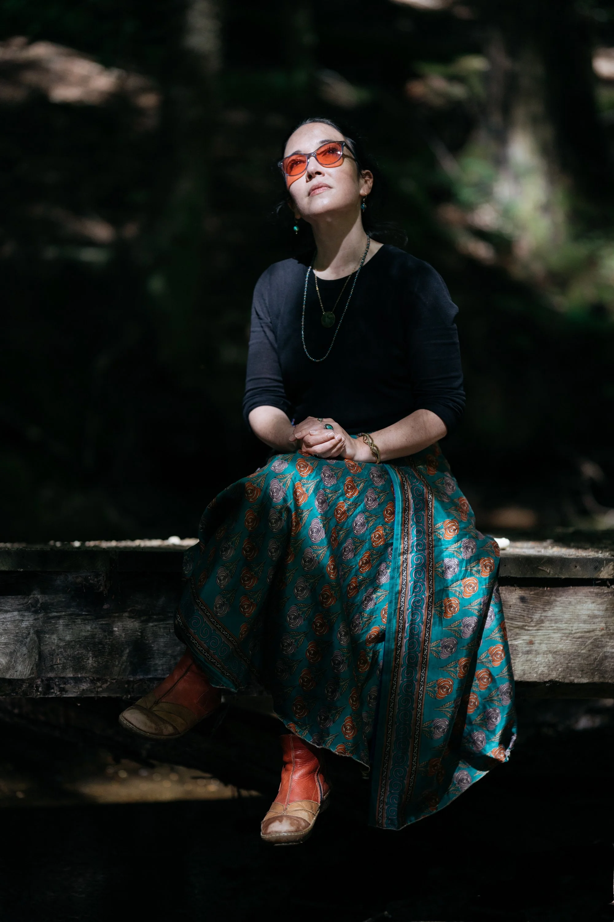 Freelance writer Jacqueline Raposo wears a long skirt, long-sleeved shirt, and migraine glasses while sitting on a bridge in the middle of the woods, her face looking up into the sky, in this photo by Paul Reitano.