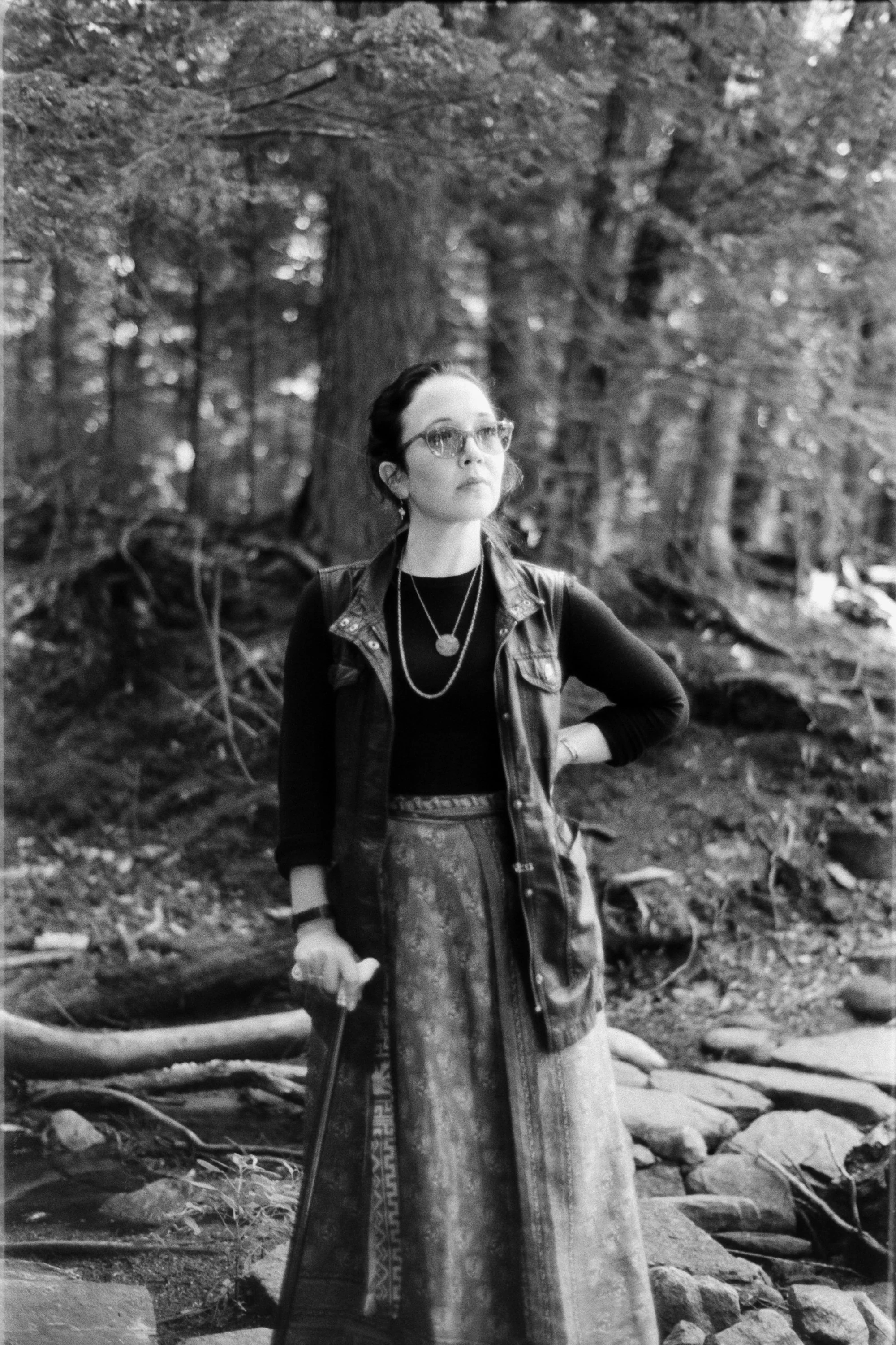 Freelance writer Jacqueline Raposo wears a long skirt, long-sleeved shirt, and migraine glasses while holding a cane and standing on a bridge in the woods in this black and white photo by Paul Reitano: