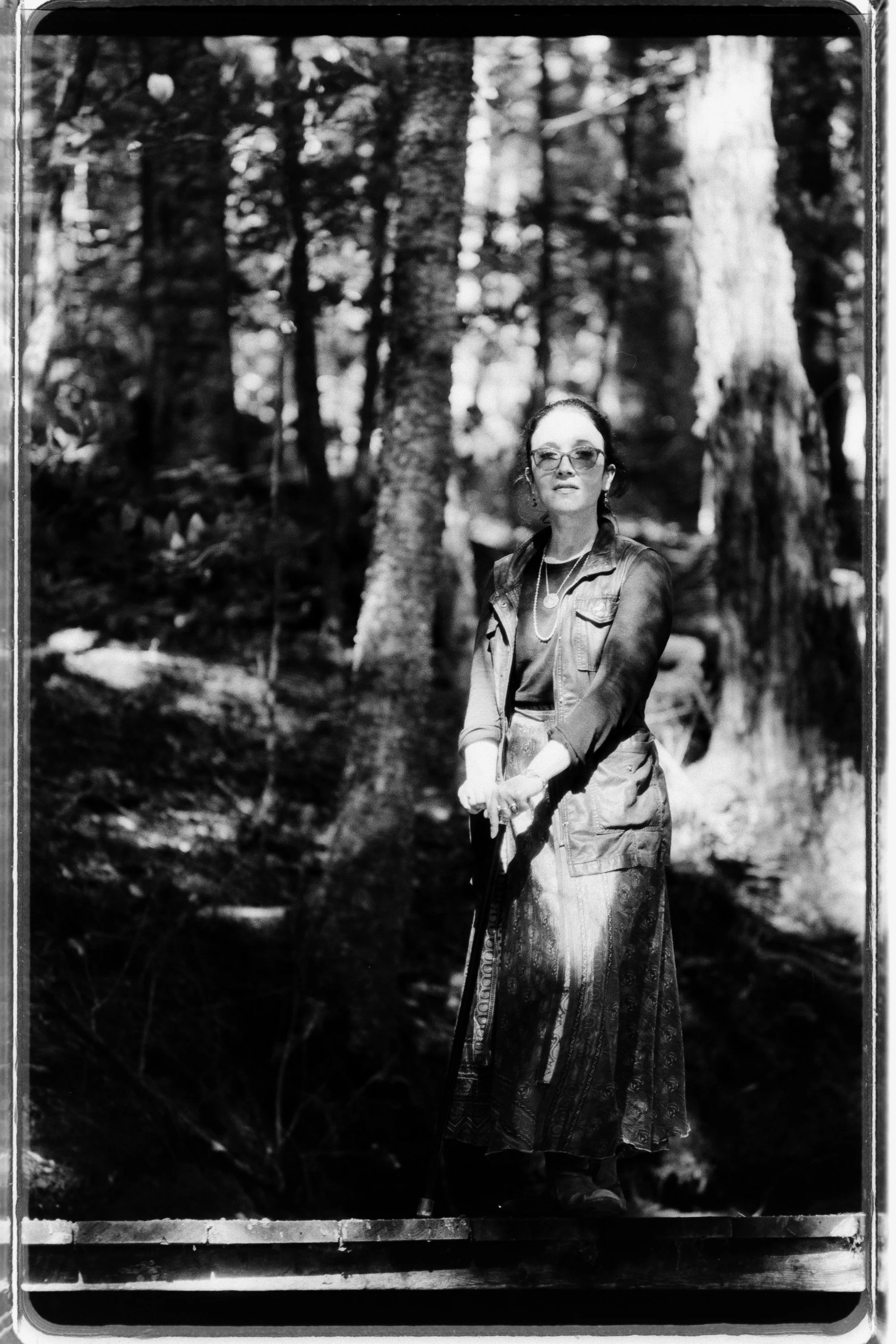 Freelance writer Jacqueline Raposo wears a long skirt, long-sleeved shirt, and migraine glasses while holding a cane and standing on a bridge in the woods looking into the camera in this black and white photo by Paul Reitano: