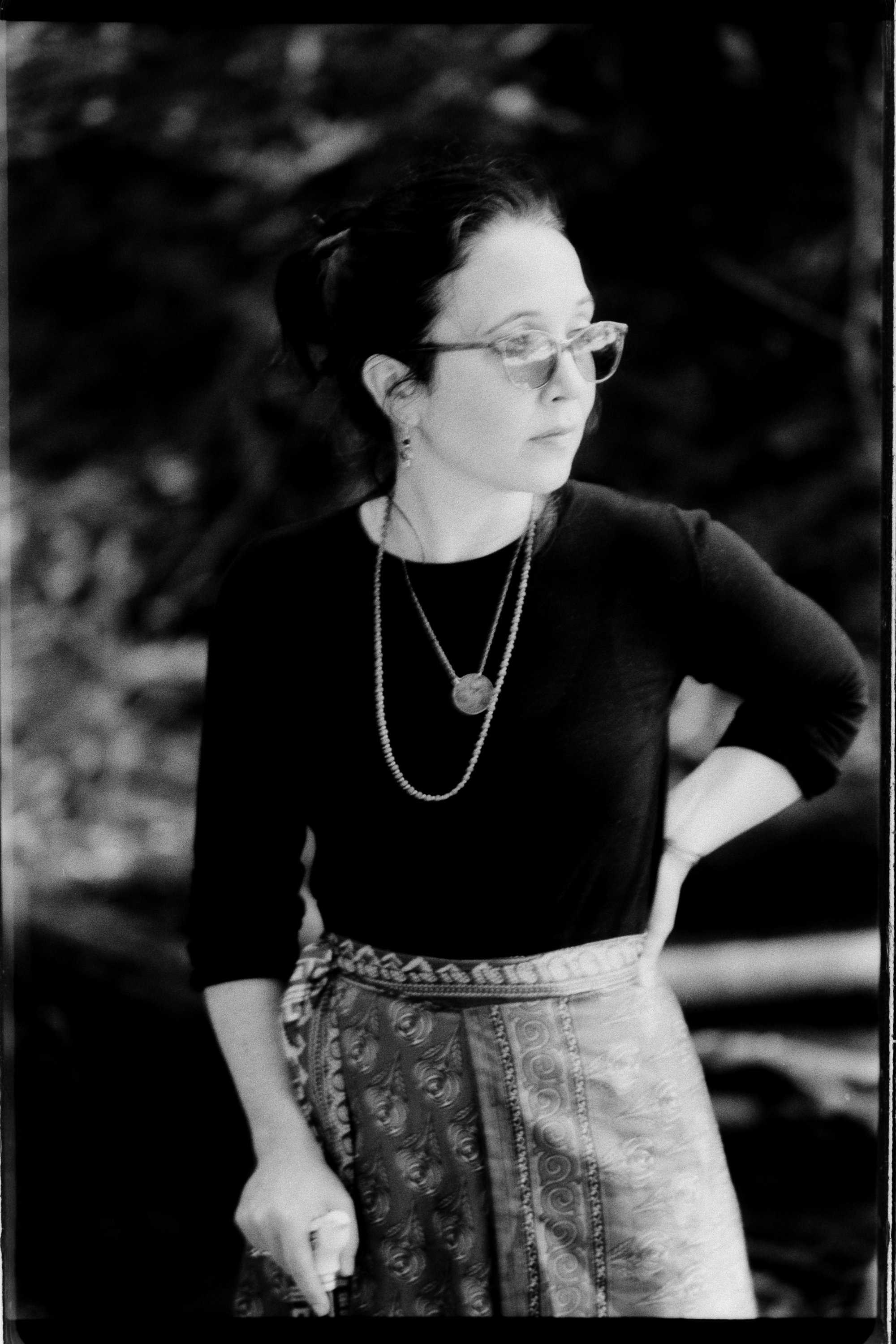 Freelance writer Jacqueline Raposo wears a long skirt, long-sleeved shirt, and migraine glasses while holding a cane and standing on a bridge in the woods in this black and white photo by Paul Reitano: