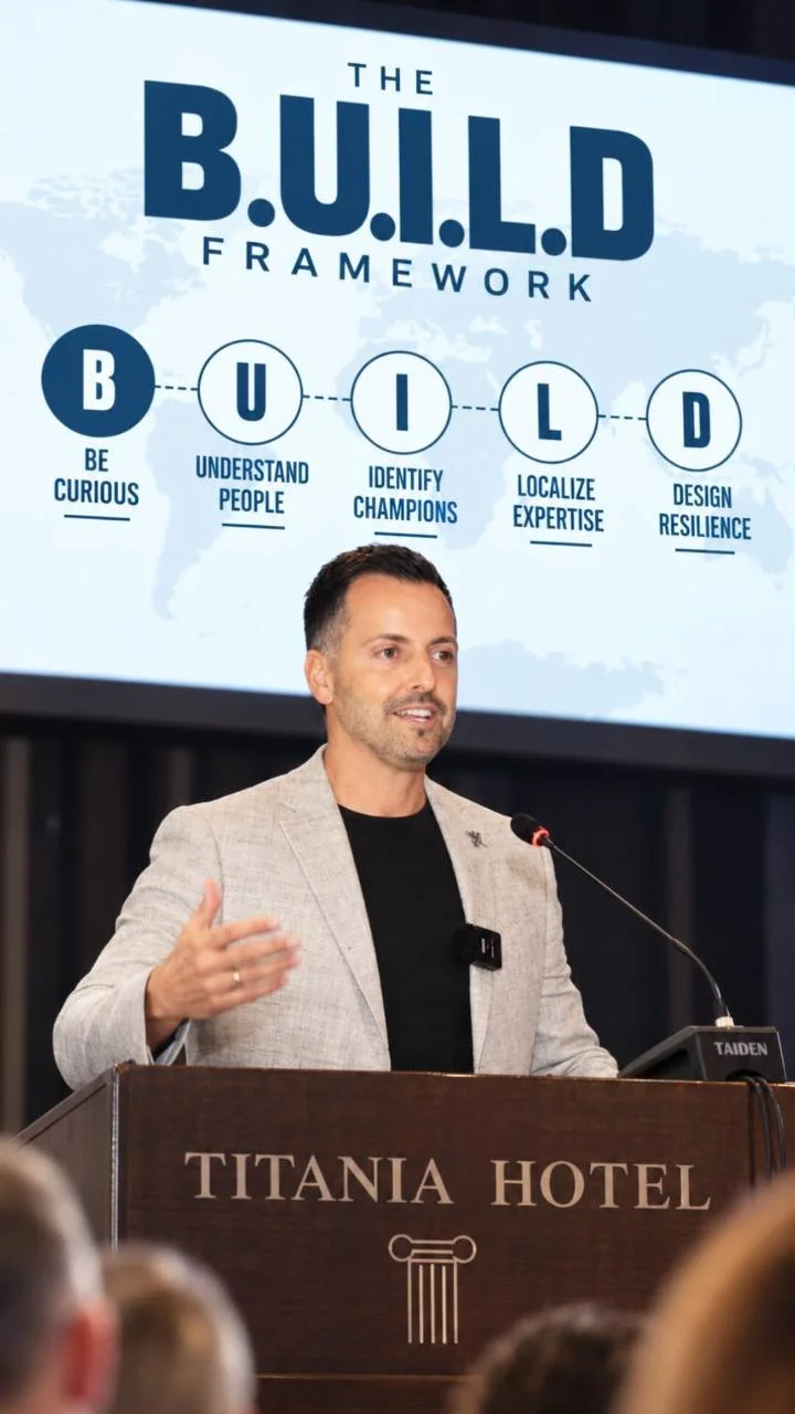 George Stroumboulis speaks at a Titania Hotel podium beneath a slide showing the five principles of his B.U.I.L.D. Framework.