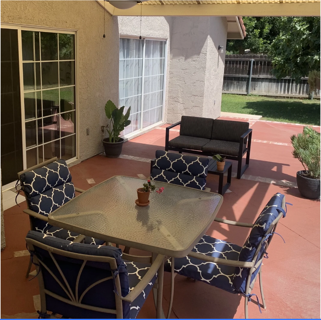 Patio with outdoor furniture, including a glass table with four chairs featuring patterned cushions, a black loveseat, a small side table with potted plants, potted plants on the patio, and a view of a grassy yard with a wooden fence.