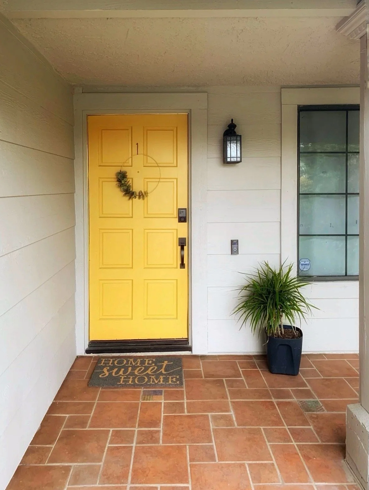 Front porch with a yellow door, a wreath, a wall-mounted lantern, a potted plant, and a doormat reading "Home sweet home."