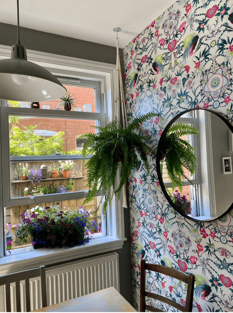 Kitchen dining nook with a wooden table and chairs, colourful floral wallpaper and a radiator beneath the window.