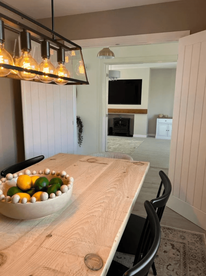 Dining room with a wooden table, black chairs and a glass lantern pendant, looking through open double doors towards a neutral living room with a stove.