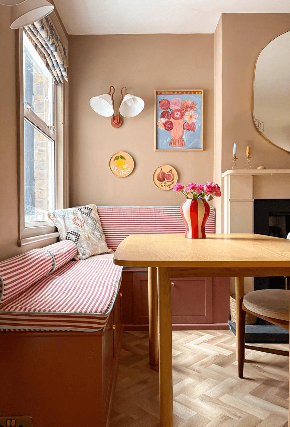 Corner bench seating with red and cream striped upholstery, a pink storage base and a wooden dining table beside a sash window.