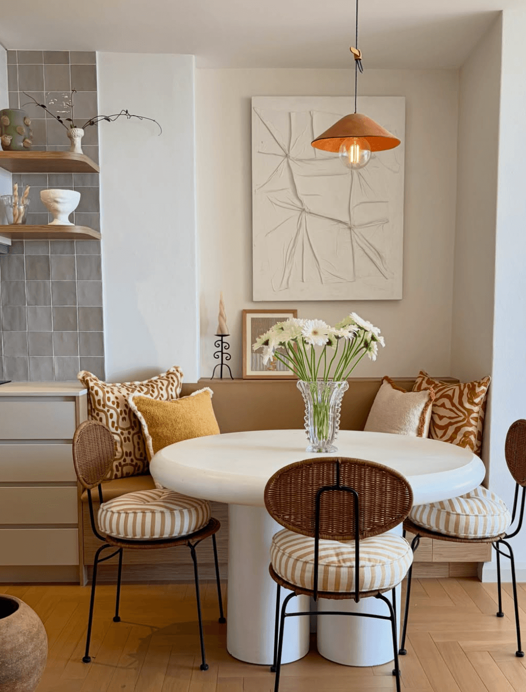 Compact kitchen dining nook with a fitted bench, patterned cushions, a white dining table and woven chairs with striped seat pads.