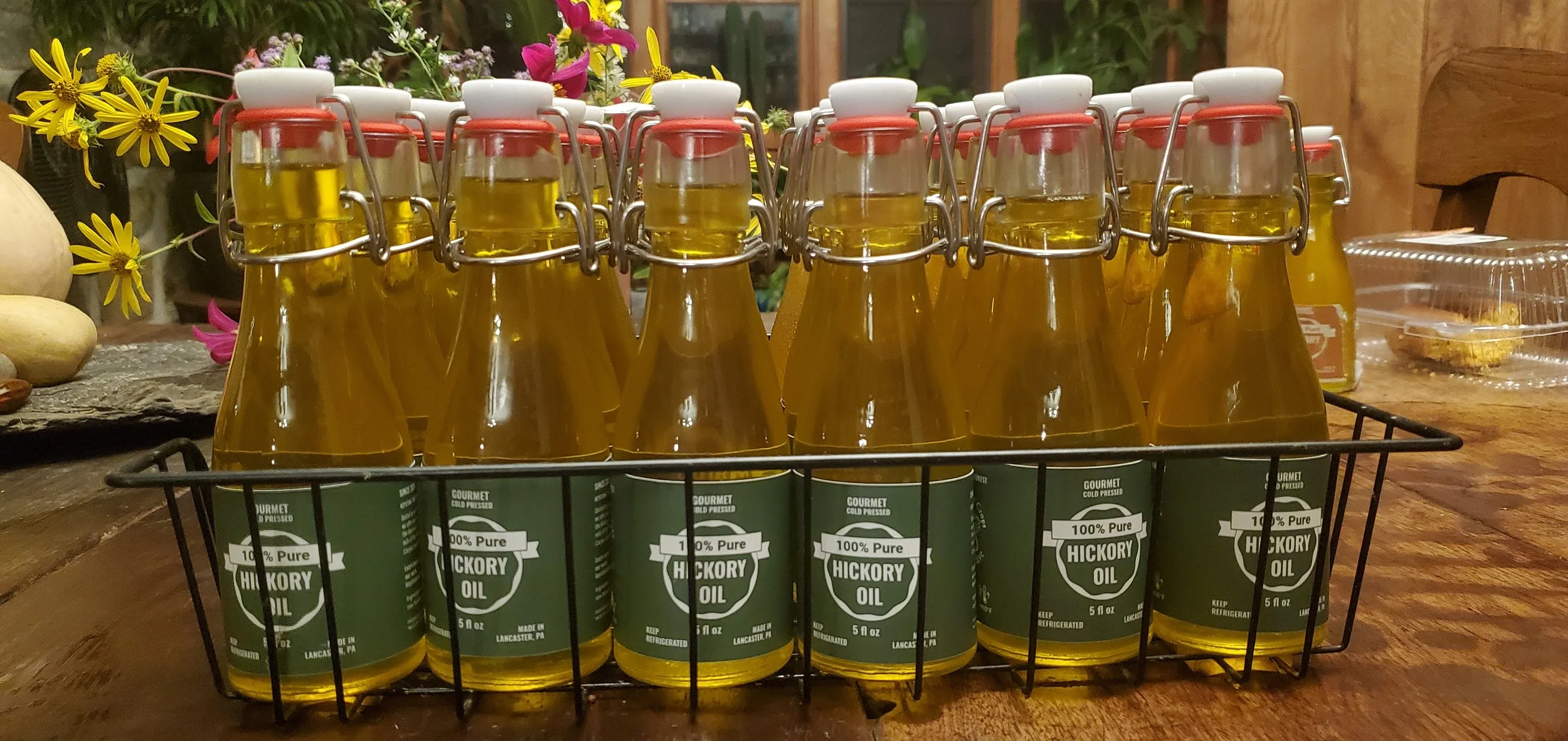 A metal basket holding multiple small glass bottles of yellowish oil labeled '100% Pure Hickory Oil' with white caps, placed on a wooden table.