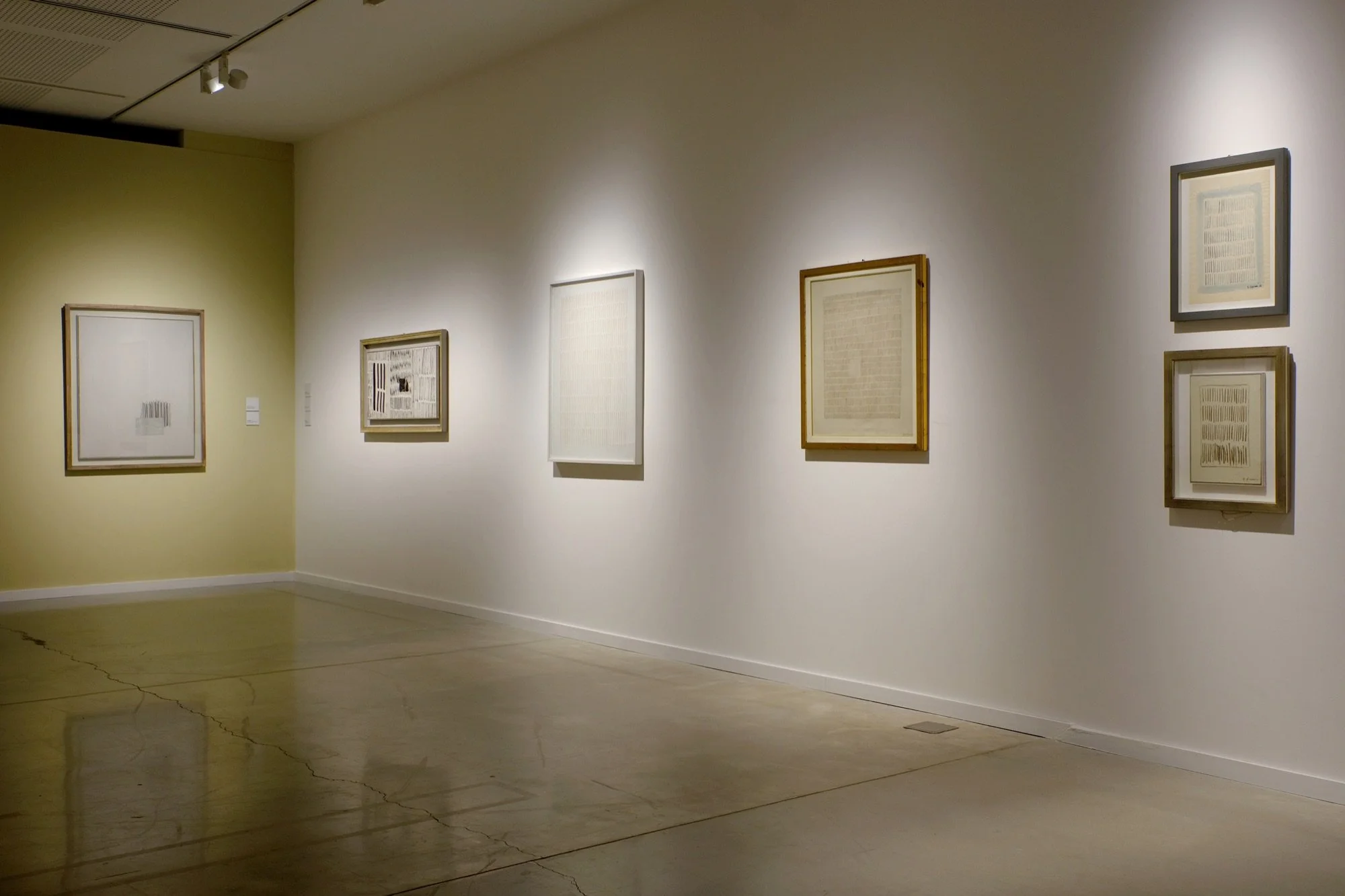 Sala de MACA con obras de Arturo Vermi enmarcadas sobre paredes blancas y una pared de color arena, iluminadas con luz cálida.