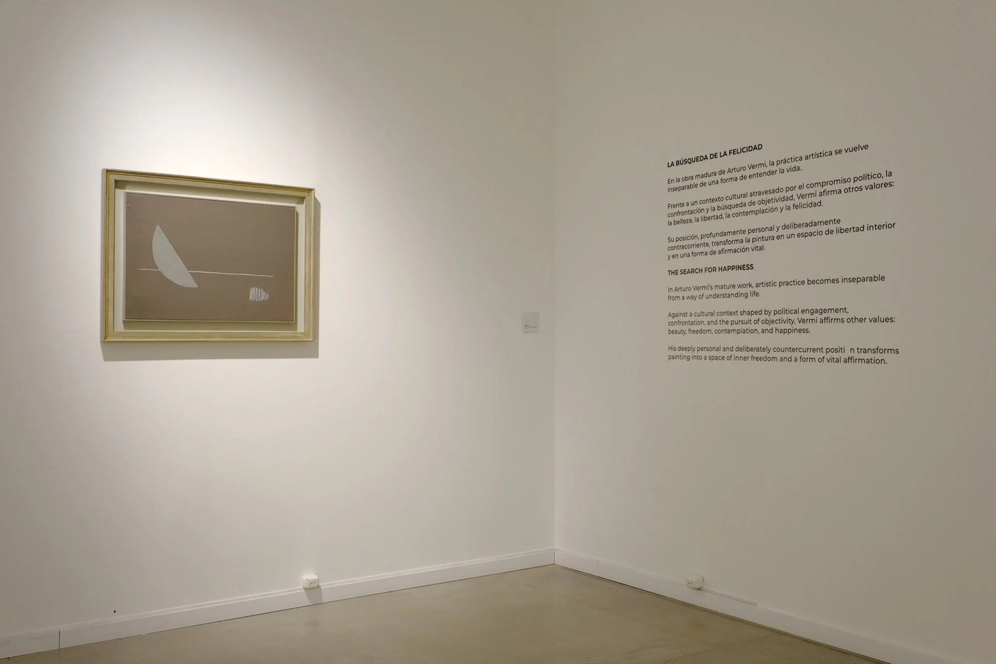 Esquina de la sala con una pintura de Arturo Vermi enmarcada y, en la pared contigua, el texto de sala «La búsqueda de la felicidad».