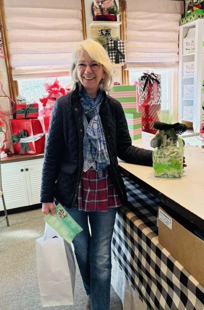 A smiling woman with glasses and blonde hair standing in a room decorated for Christmas, holding a gift wrapped in green and white paper with a black bow. Behind her are shelves with holiday decorations and a table with more wrapped presents, surroun
