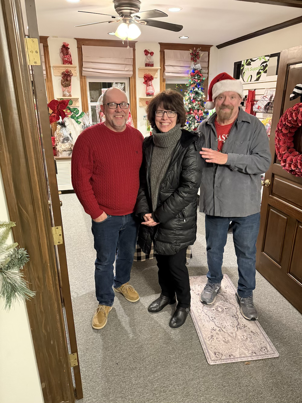 Three people standing in a room decorated for Christmas with a Christmas tree, wreaths, and ornaments. One man is wearing a red sweater, a woman in the middle is wearing a gray turtleneck and black coat, and the man on the right is wearing a Santa ha