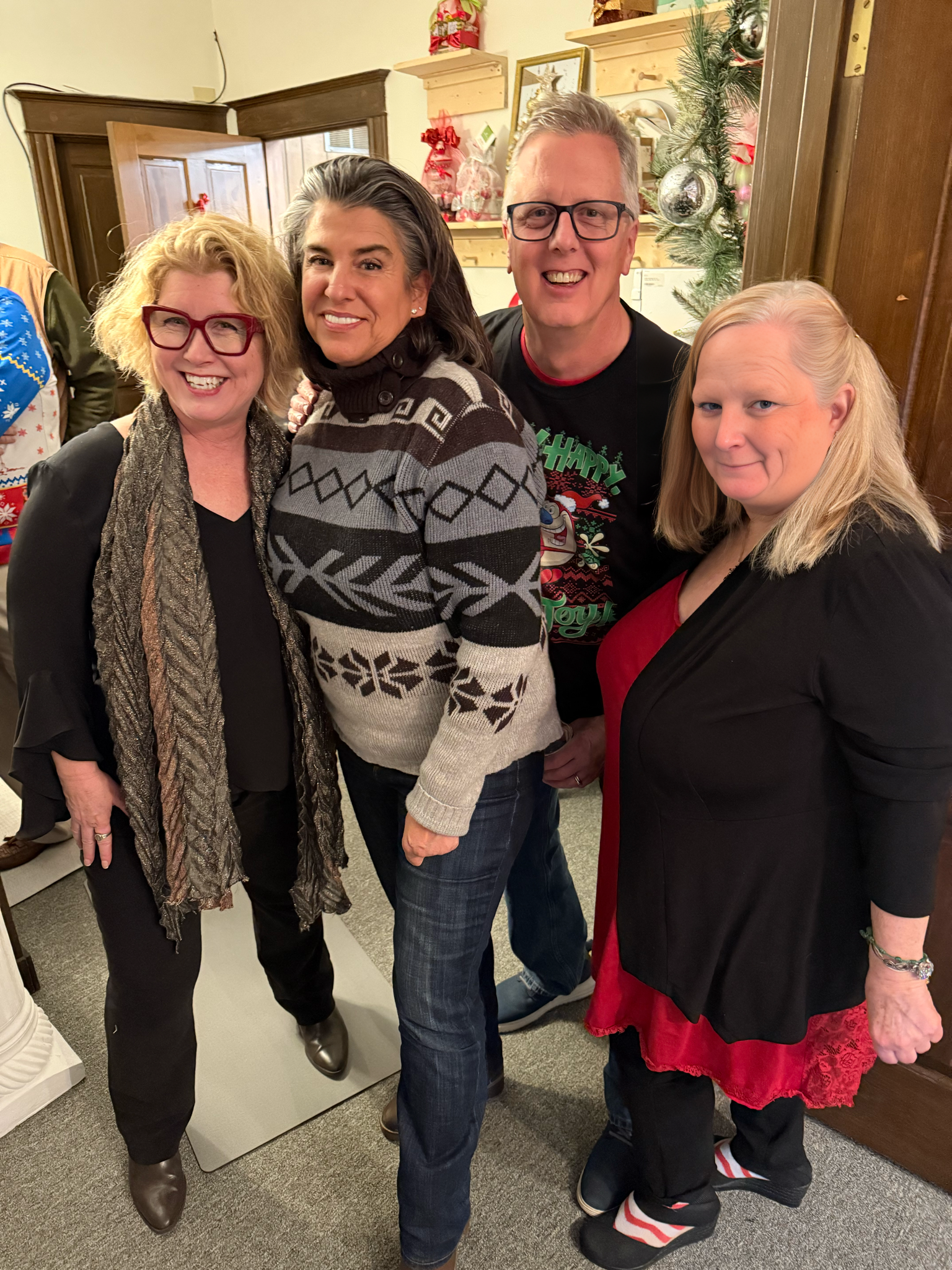 A group of four smiling adults posing indoors near Christmas decorations, including a Christmas tree, in a festive setting.