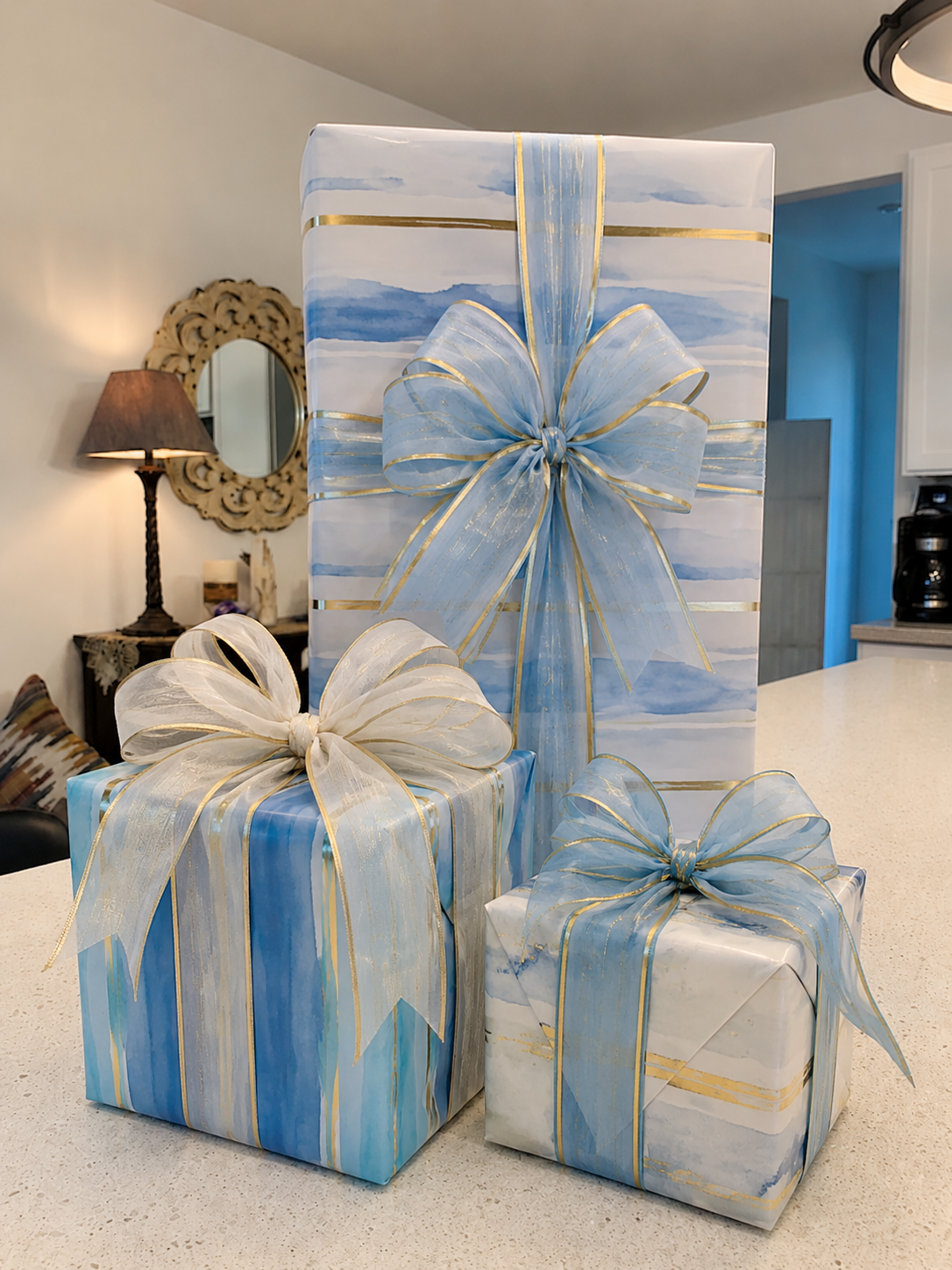 Three wrapped gift boxes with blue and white marbled wrapping paper and large, sheer bows with gold accents, placed on a white countertop in a home.