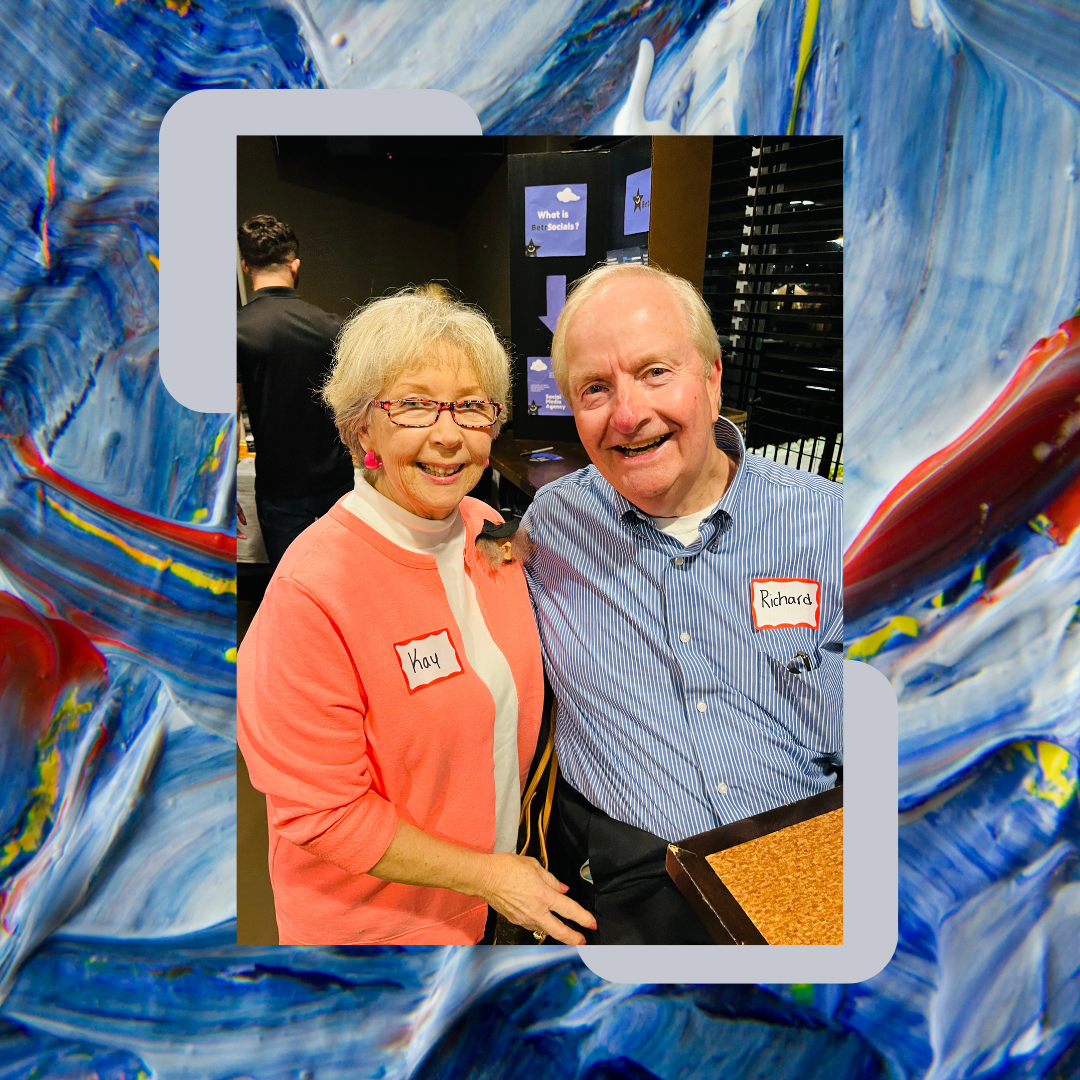 A smiling senior woman wearing glasses, earrings, and a pink cardigan with a white shirt underneath, standing next to a smiling senior man with a name tag that says 'Richard,' at a social event. Both are looking at the camera. The woman has a label t