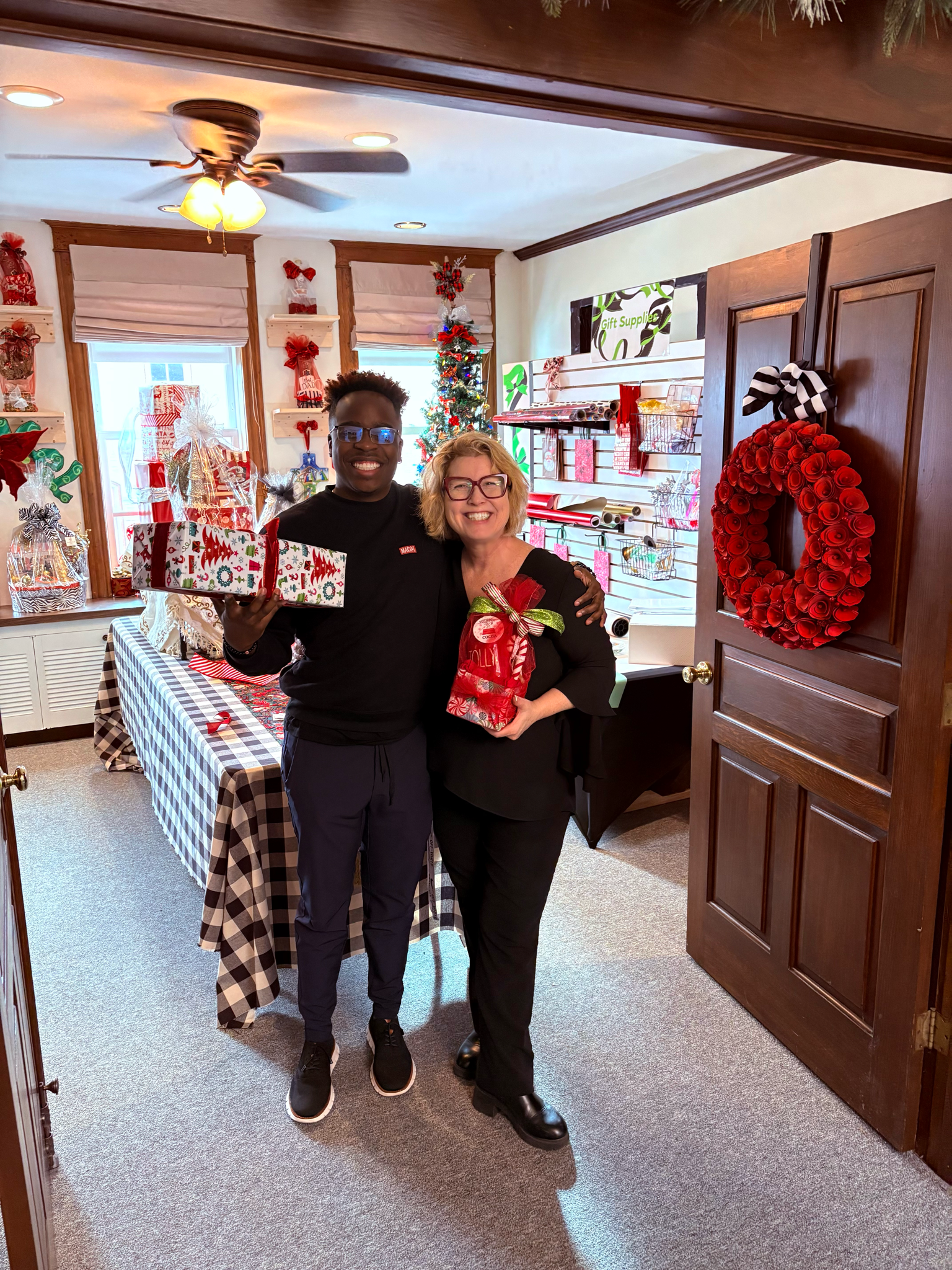 Two women smiling and holding Christmas presents inside a decorated room with holiday ornaments, a Christmas tree, and a checkered tablecloth.