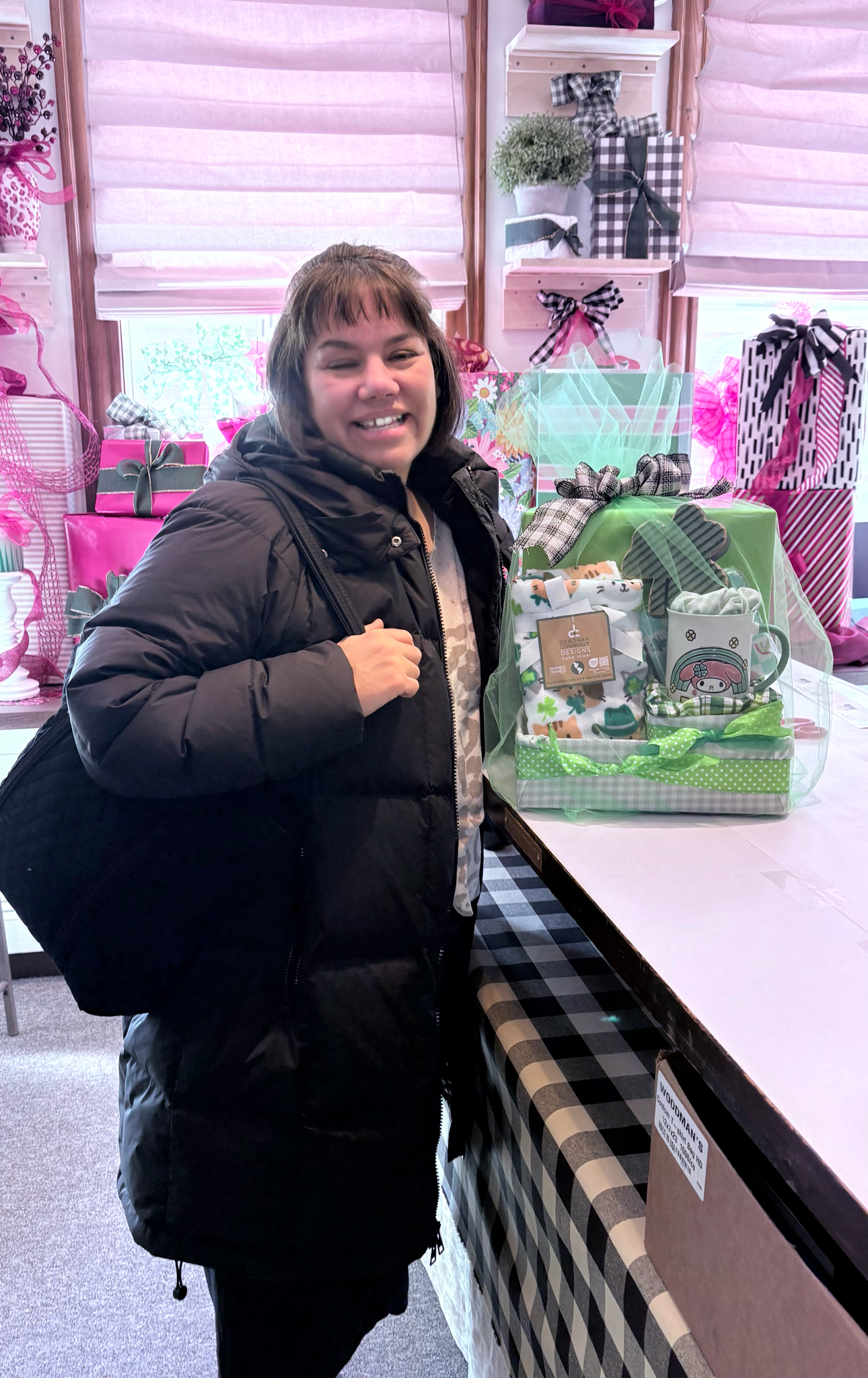 Woman smiling next to a gift basket with a mug, socks, and other wrapped items, in a store with pink and black plaid decorations.