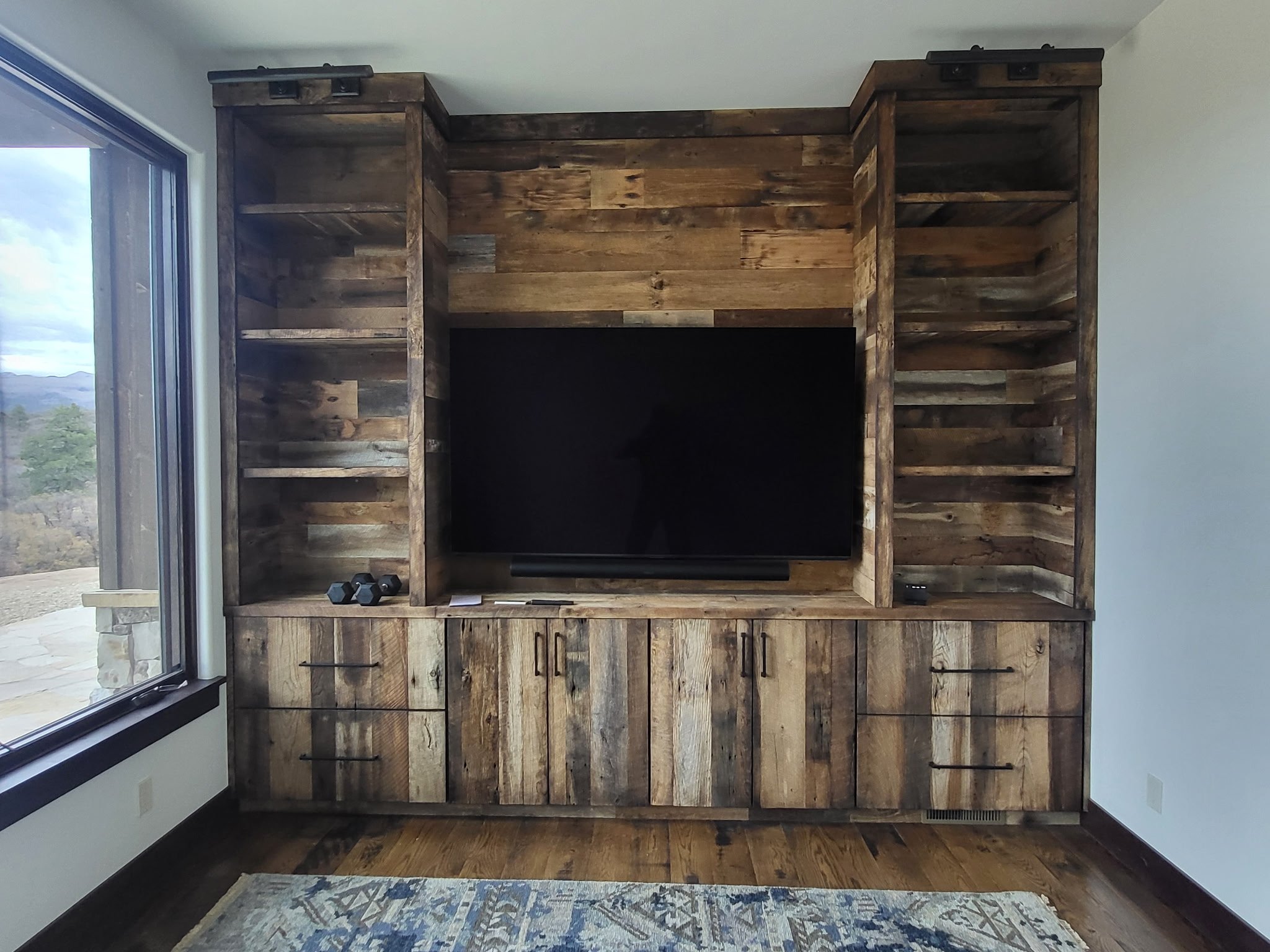 Custom reclaimed wood media center with central TV space, two bookcase towers, full-width base cabinetry, matching reclaimed wood countertop, and crown molding to ceiling—modern functionality with rustic style.